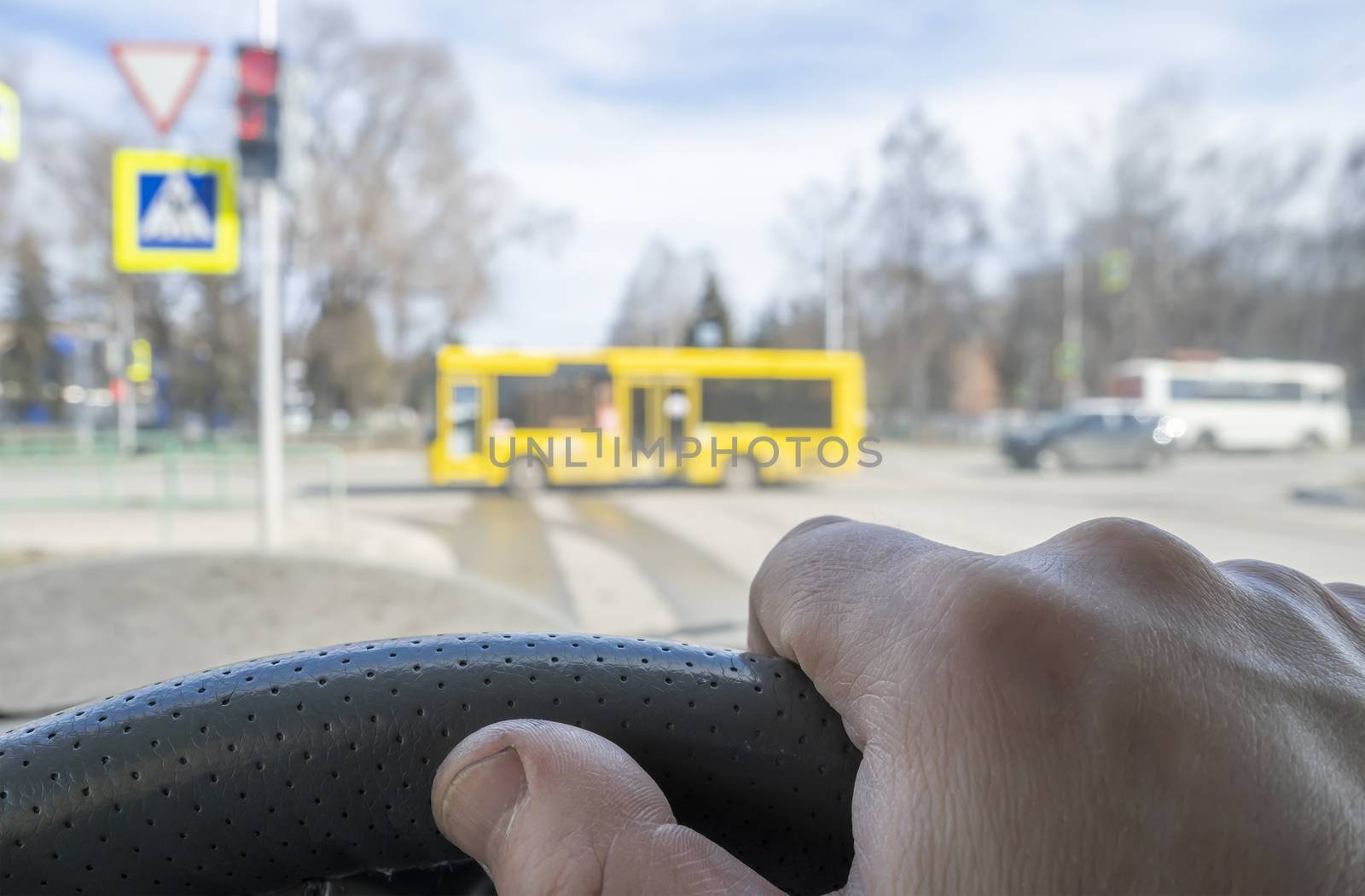 the driver hand on the steering wheel of a car located in front of a red prohibiting traffic light, pedestrian crosswalk, and cars that the driver gives way to
