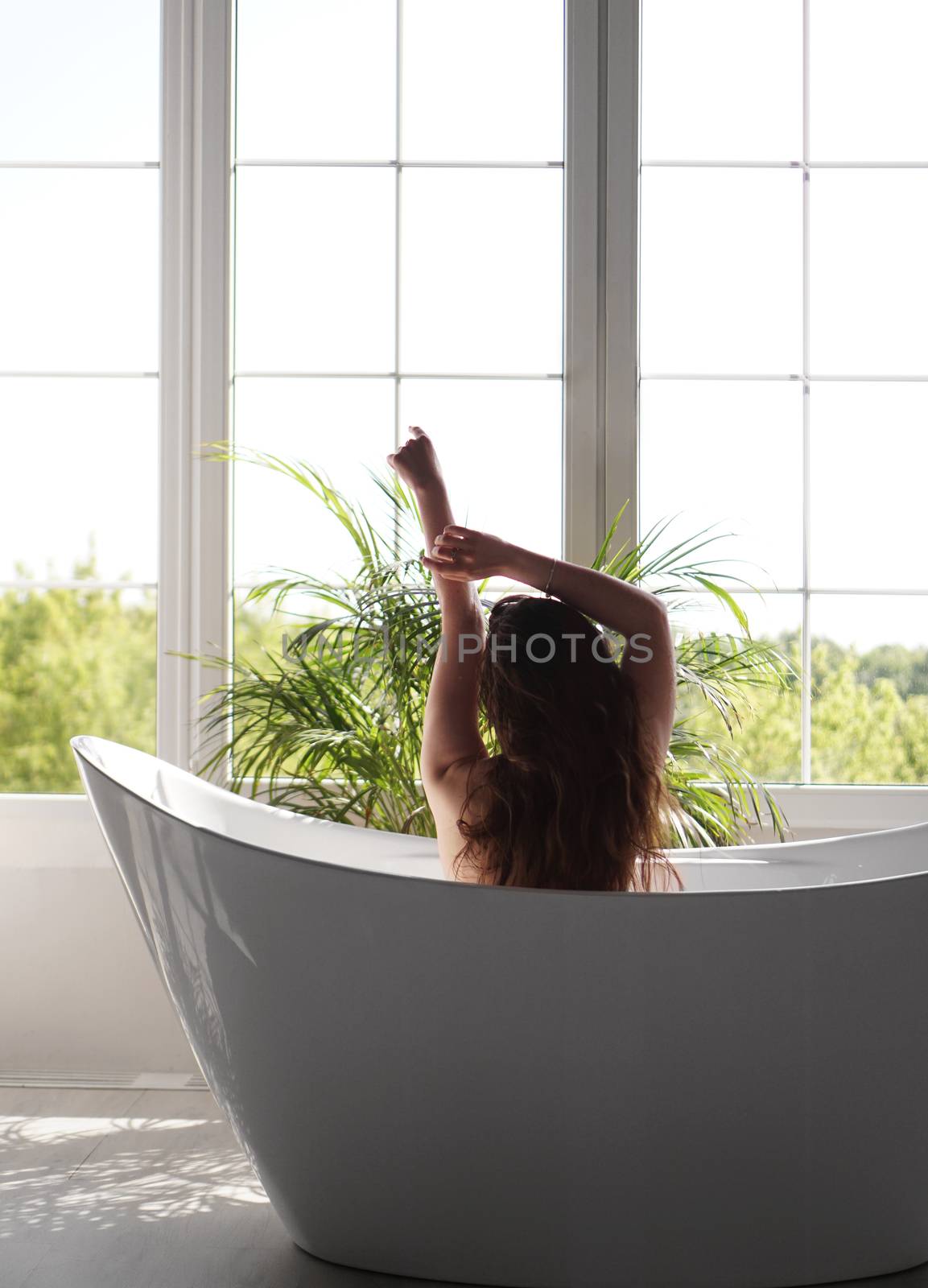 Backside portrait of fit slim woman against the window in bathroom. Green plant in bathroom
