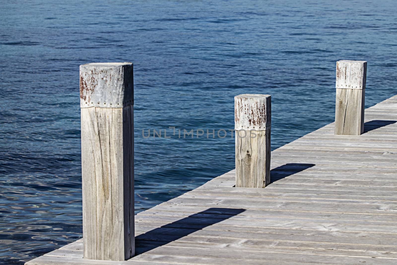 A small quay and its three square mooring poles that create geometric games with the shadows and the symmetry of the wooden planks of the quay on the blue sea