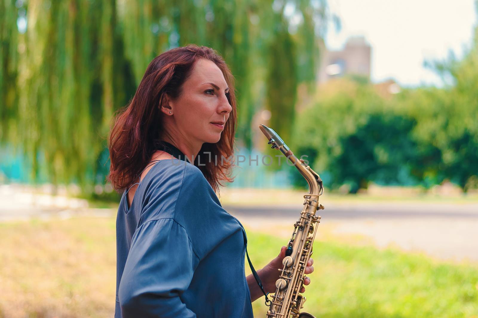 cute young redhead girl in a blue blouse posing with a saxophone in the park by chernobrovin