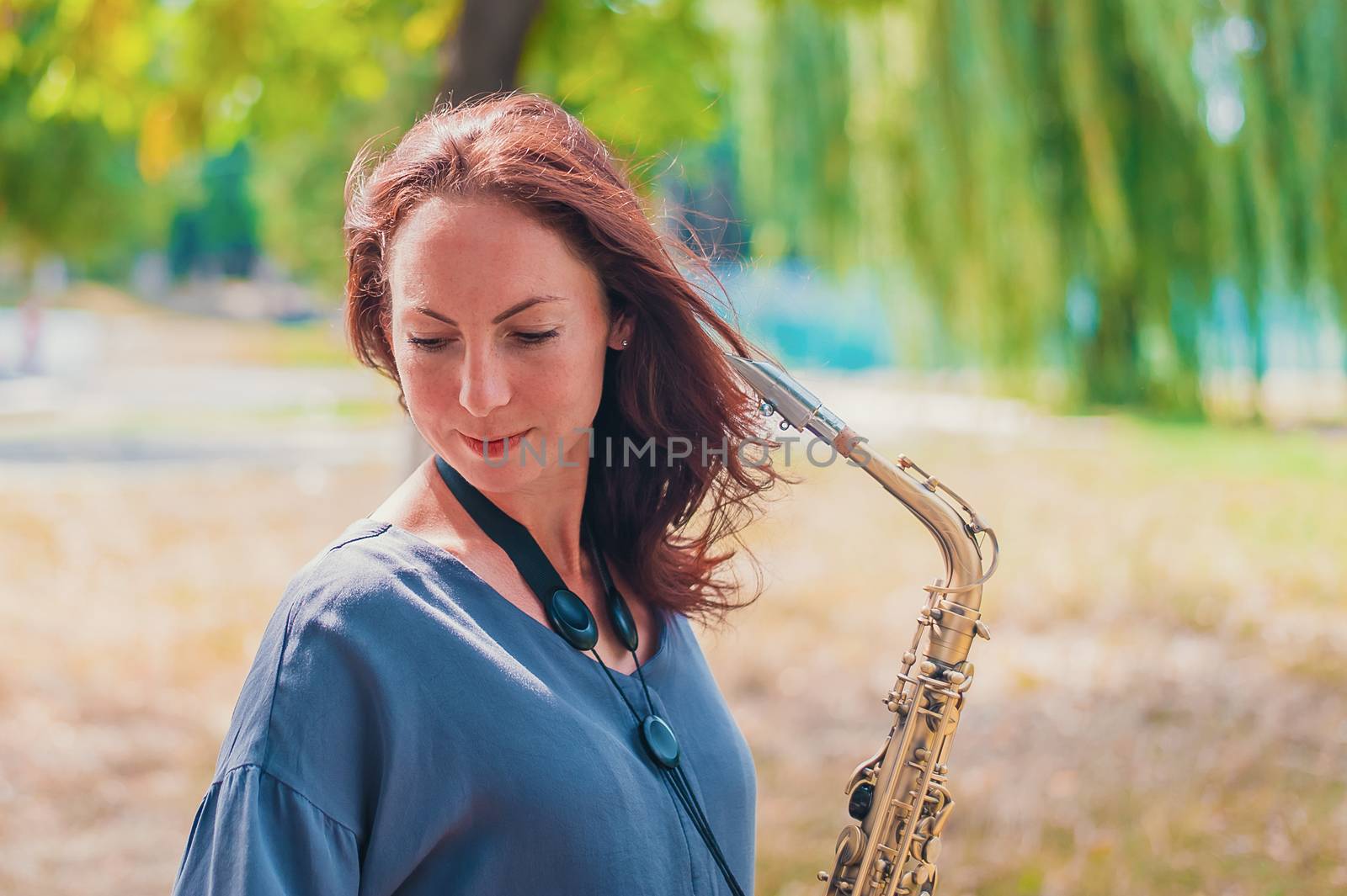 Close portrait of a young red-haired woman in a green park with a saxophone in summer by chernobrovin