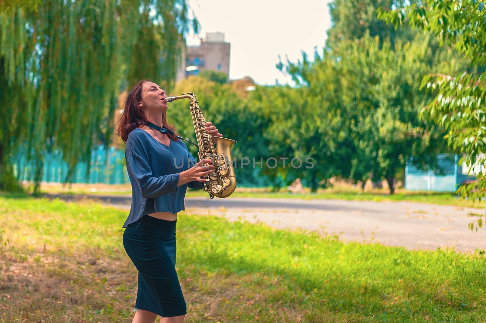 pretty young woman with red hair in a blue sweater and black skirt plays on a yellow saxophone in a city green park by chernobrovin