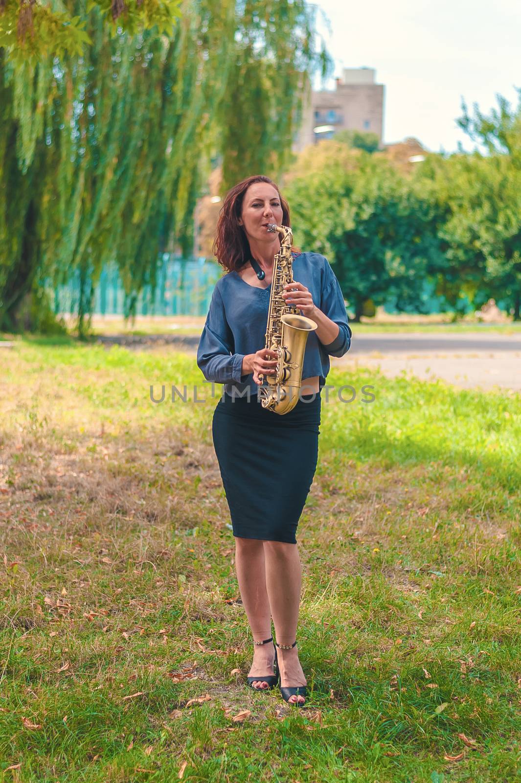 nice girl with red hair in a blue sweater and black skirt plays on the saxophone in the park