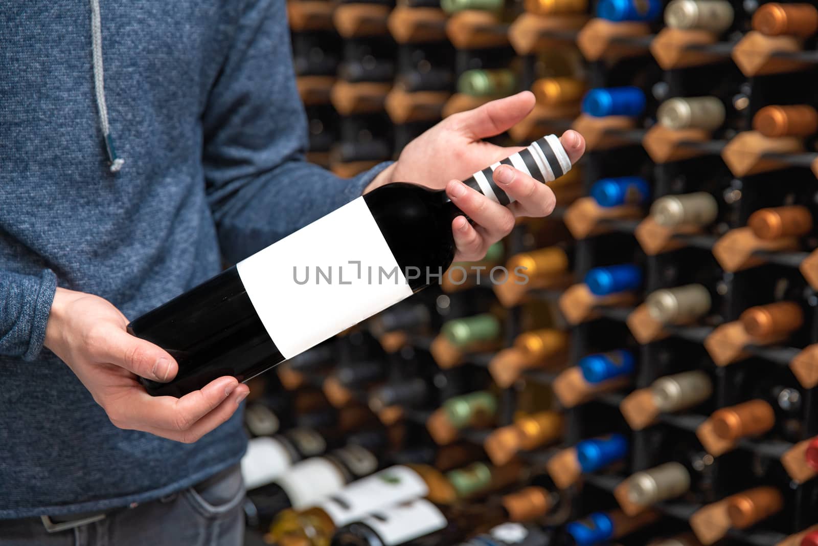 man in a wine shop holding a bottle with a blank label in his hand. A rack with wine in the background.