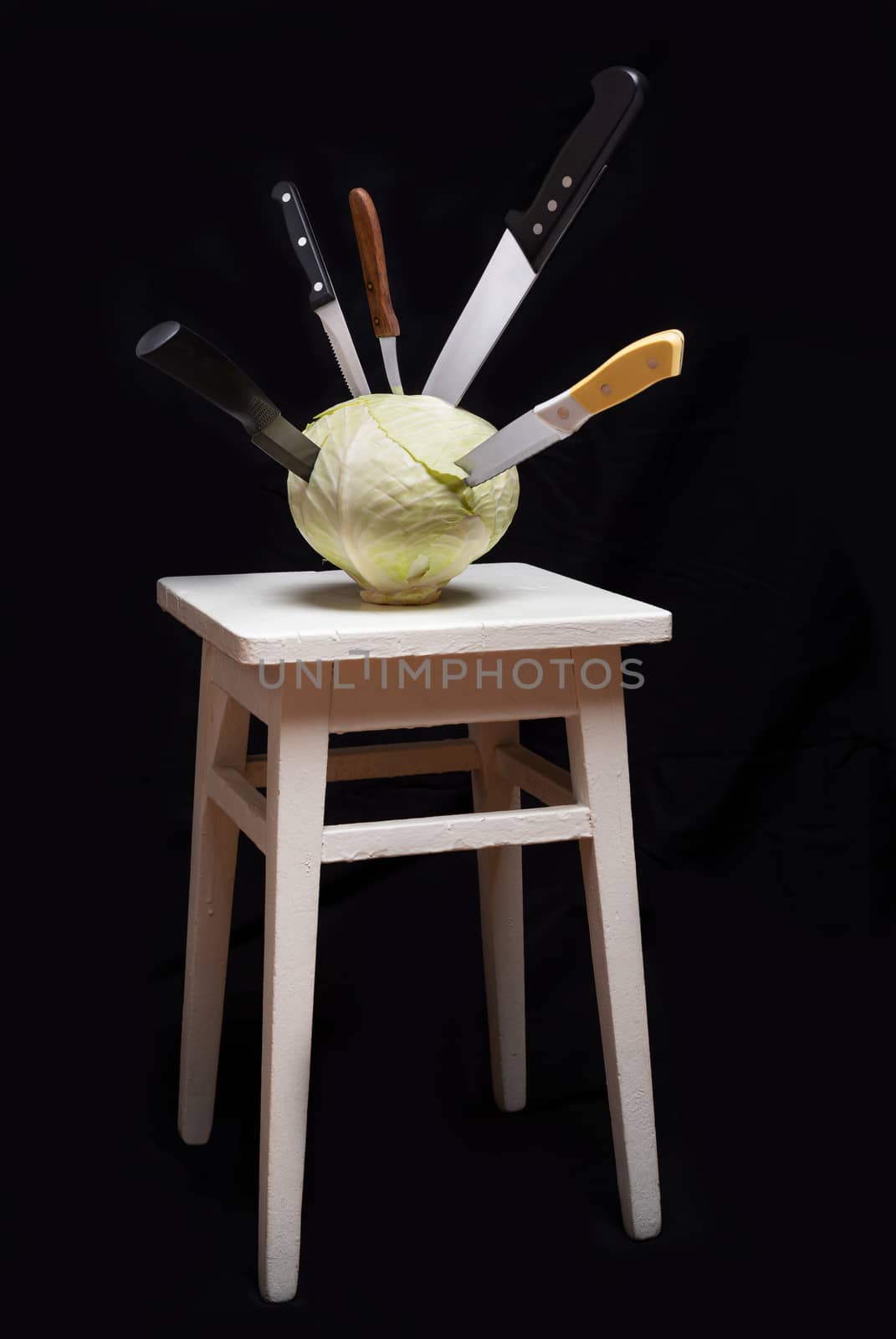 Symbolic conceptual image of a white cabbage on a white wooden stool. Knives are stuck into the vegetable and symbolize cooking, violence, crime, suffering. On black background.