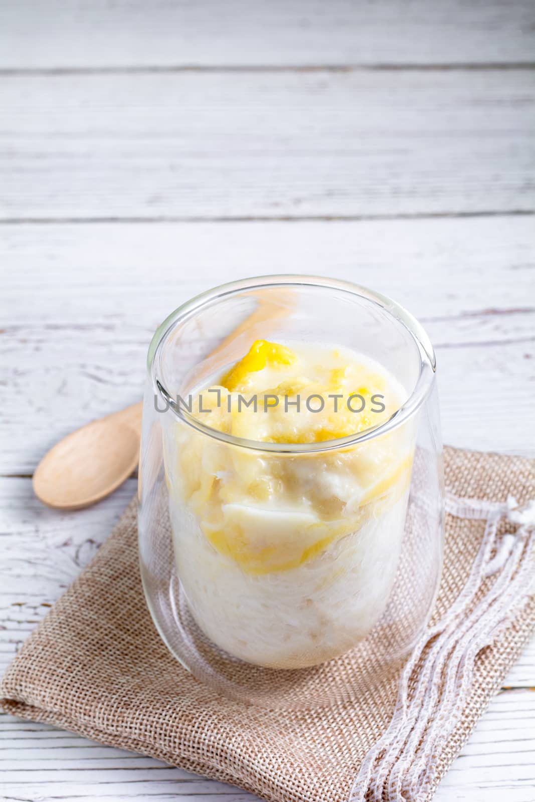 Summer appetizer of Thailand concept. Durian and sticky rice with coconut milk cream in transparent glass on white wooden table and brown table cloth. Famous street food in Thailand.  