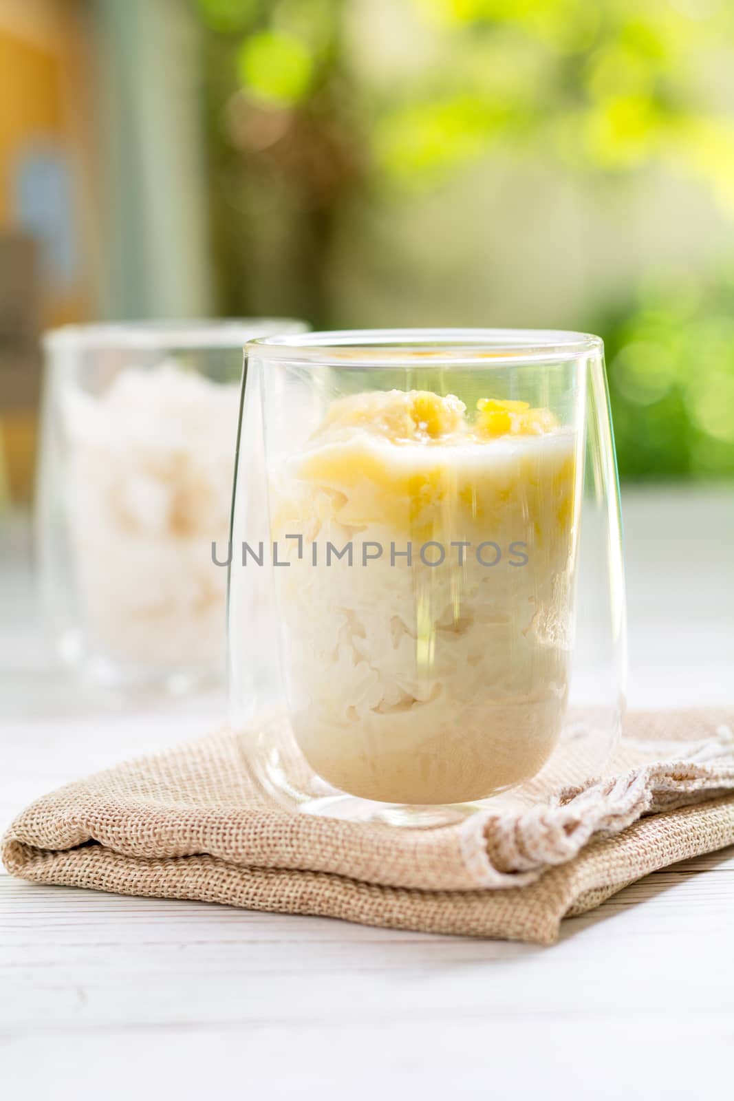Summer appetizer of Thailand concept. Durian and sticky rice with coconut milk cream in transparent glass on white wooden table and brown table cloth and yellow and green bokeh light background. Famous street food in Thailand.  