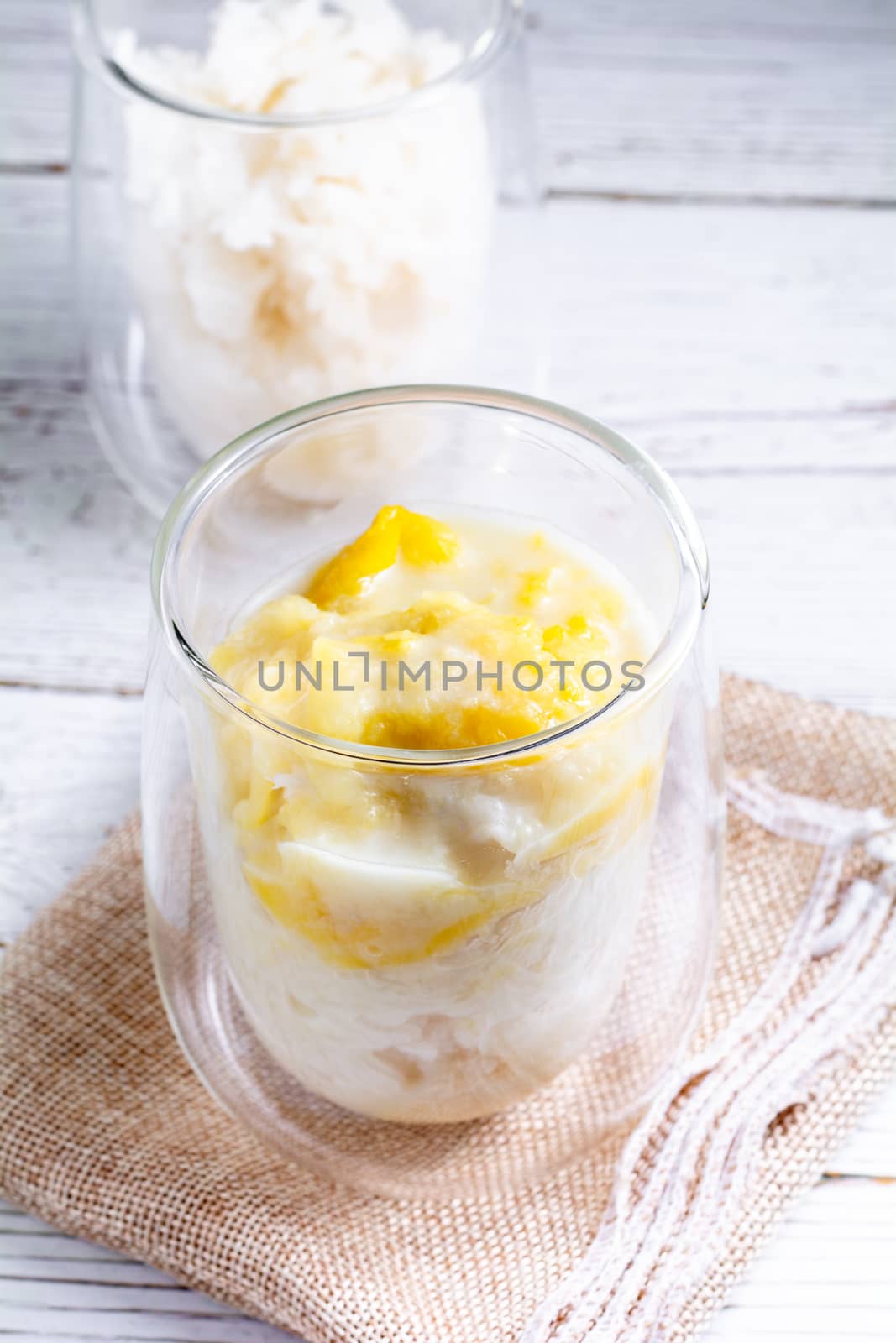 Summer appetizer of Thailand concept. Durian and sticky rice with coconut milk cream in transparent glass on white wooden table and brown table cloth. Famous street food in Thailand.  