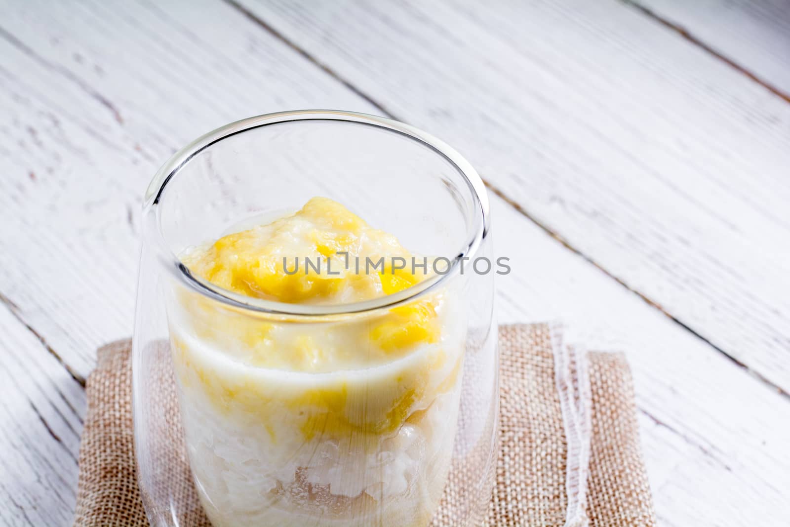 Summer appetizer of Thailand concept. Durian and sticky rice with coconut milk cream in transparent glass on white wooden table and brown table cloth. Famous street food in Thailand.  