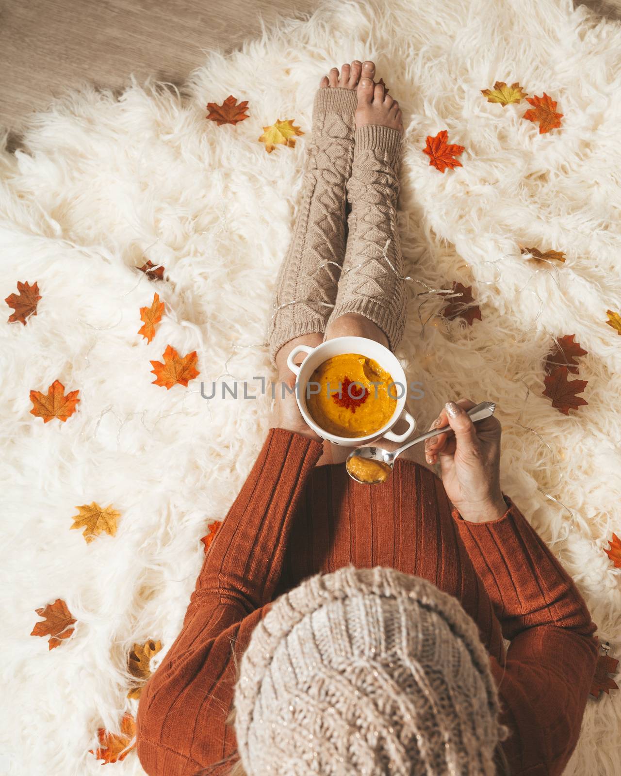 Woman on a fluffy cozy rug holds a bowl of spiced pumpkin soup in the fall autumn season. Overhead perspective with scattered maple leaves