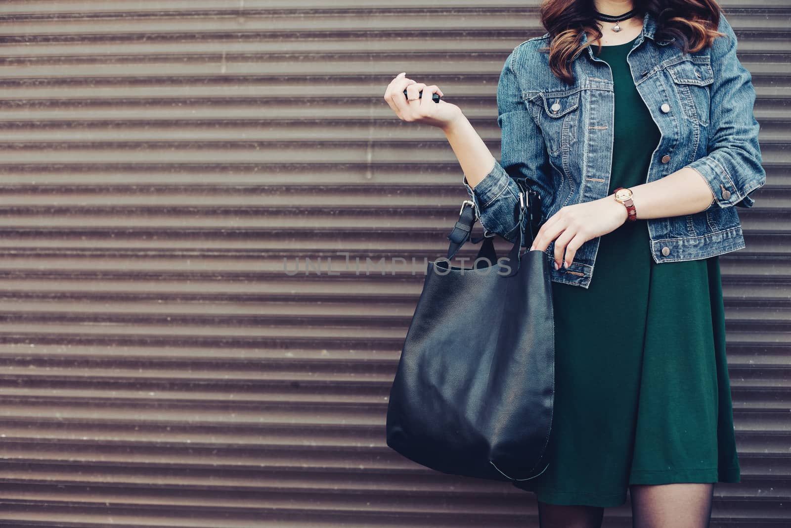 Crop of stylish incognito girl with big black leather bag after shopping, posing at street, leaning on wall. Woman in dress, jeans jacket, with accessories. Street fashion.