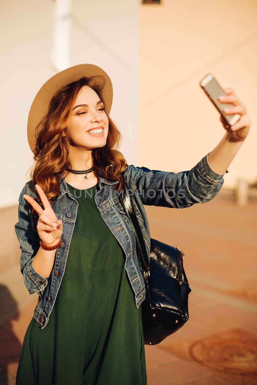 Brunette girl in sunglasses and hat making duck face, sending kiss at camera and taking self portrait at smart phone.Beautiful and funny woman posing, walking at street. Concept of street fashion.