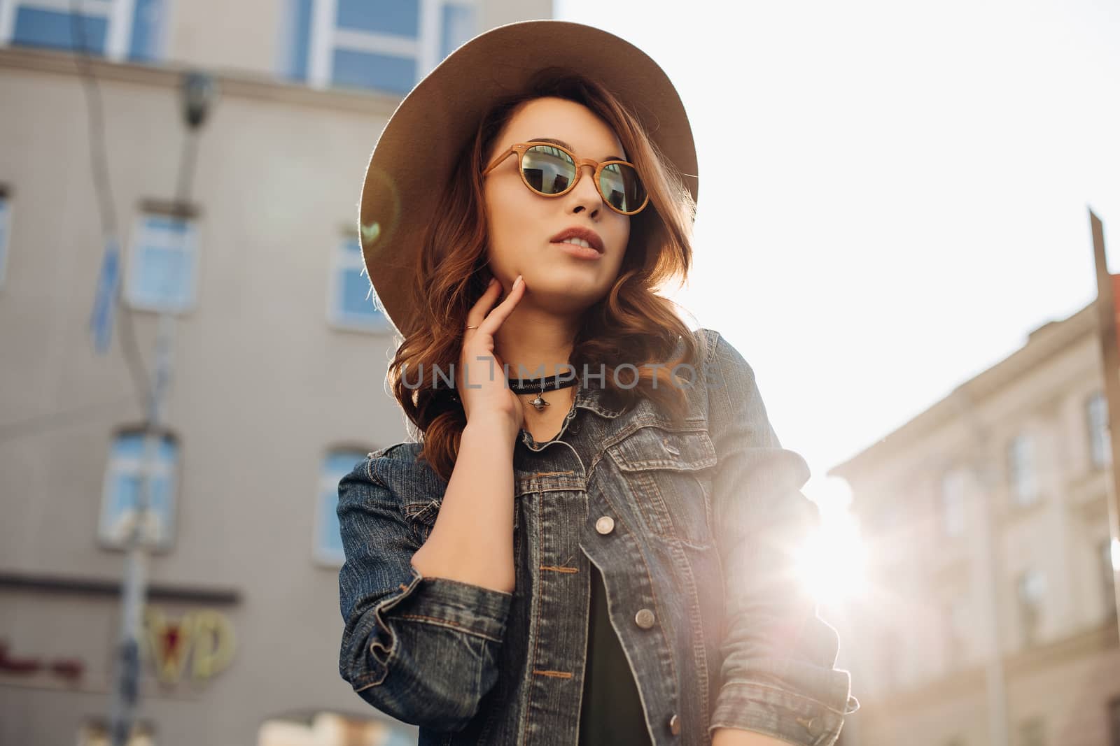 Portrait of beautiful brunette woman stylish wearing in hat, choker, jeans jacket holding glasses in hand and posing. Attractive girl walking at street, smiling at camera. Street fashion and beauty.