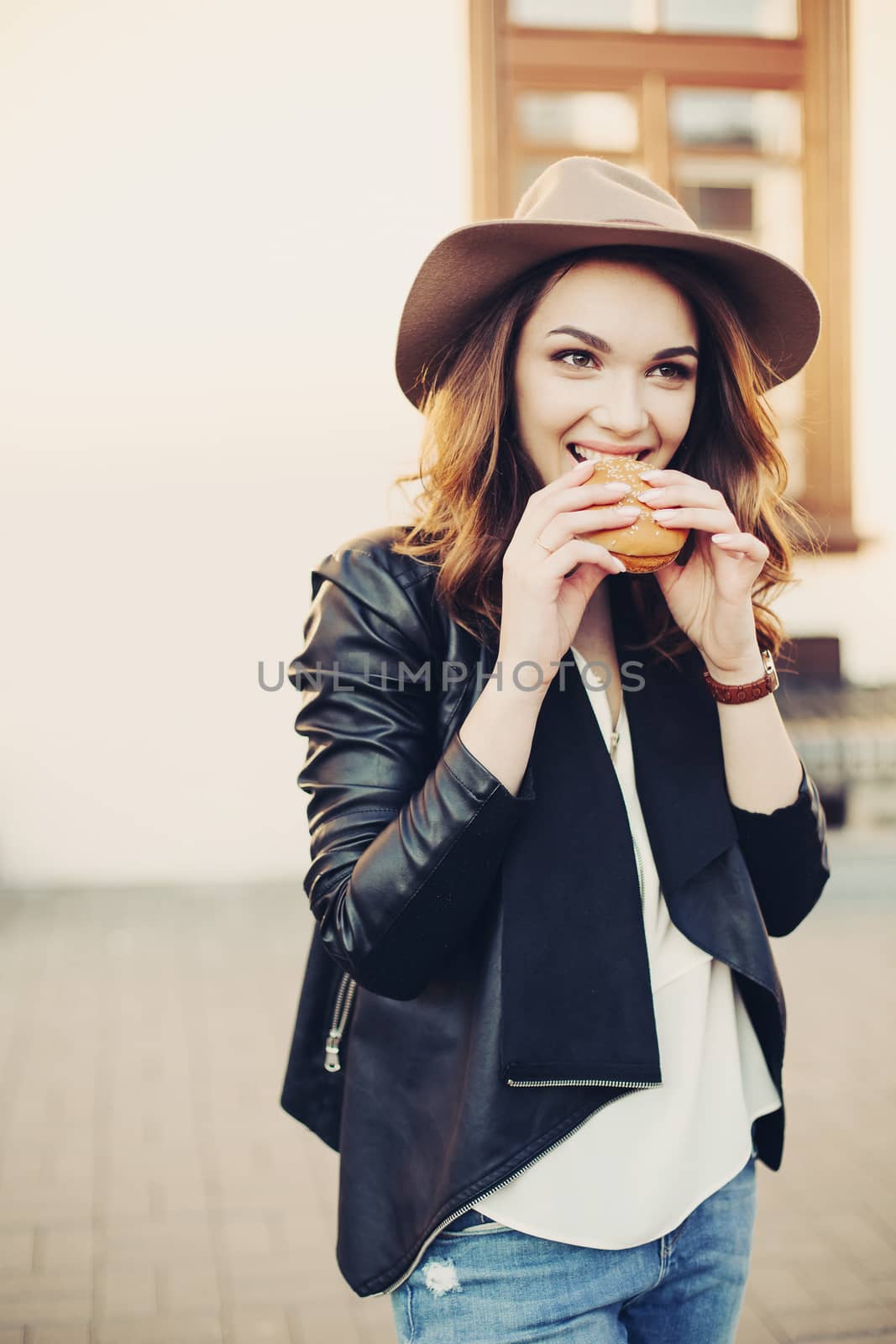 Beautiful and fashionable brunette woman in brown hat eating tasty hamburger with clossed eyes and enjoying. Emotionally girl having dinner of fast food at street, sensuality smiling.