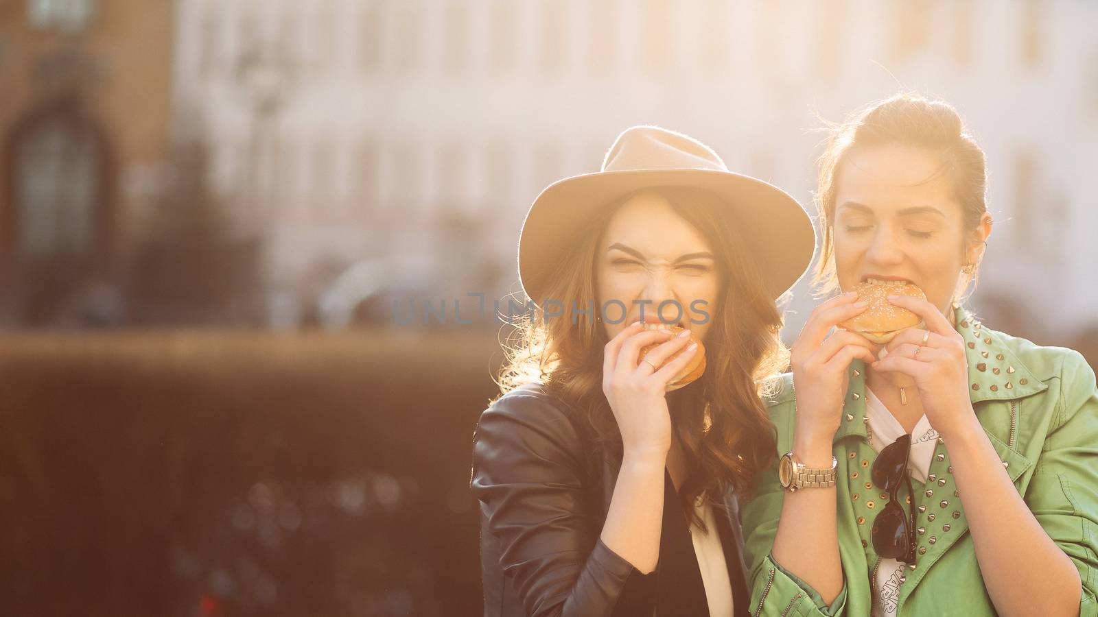 Positivity girls eating fast food, hamburgers, having dinner together, smiling at camera and posing. Beautiful couple of stylish girlfriends in leather jackets and hat, eating junk and unhealthy food.