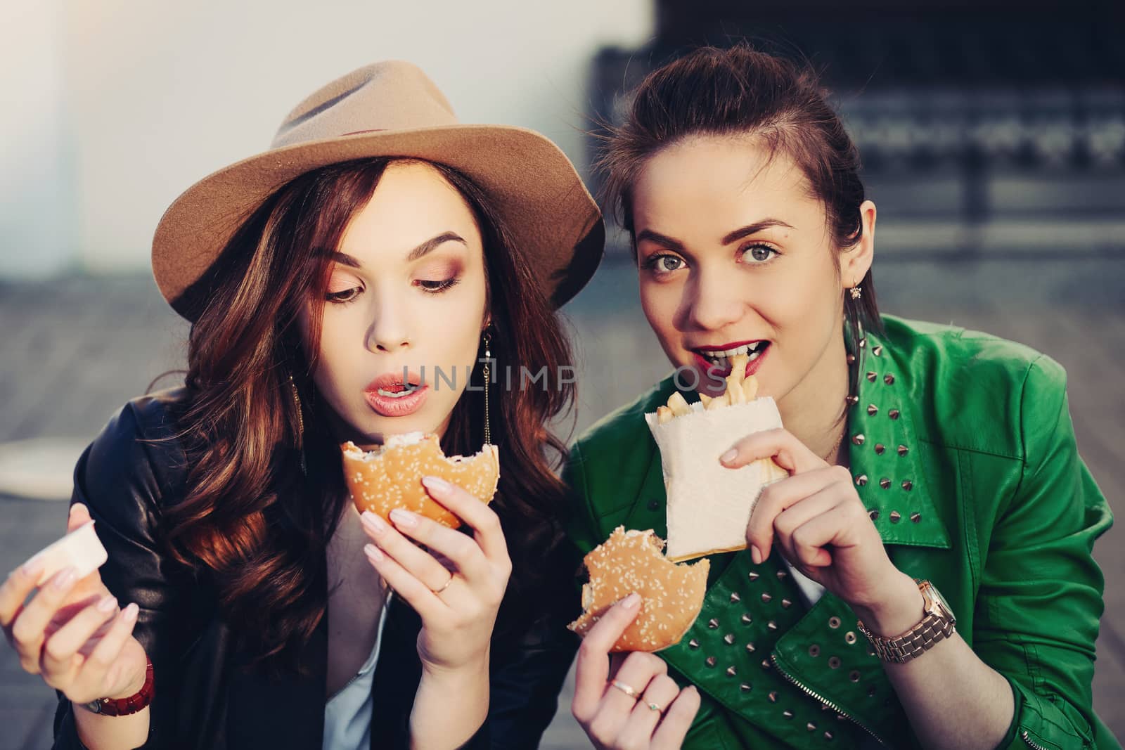 Funny girls sitting at street and eating fast food, having dinner together, and smiling to each other. Beautiful young girlfriends posing with hamburger and potato fried. Junk and unhealthy food.