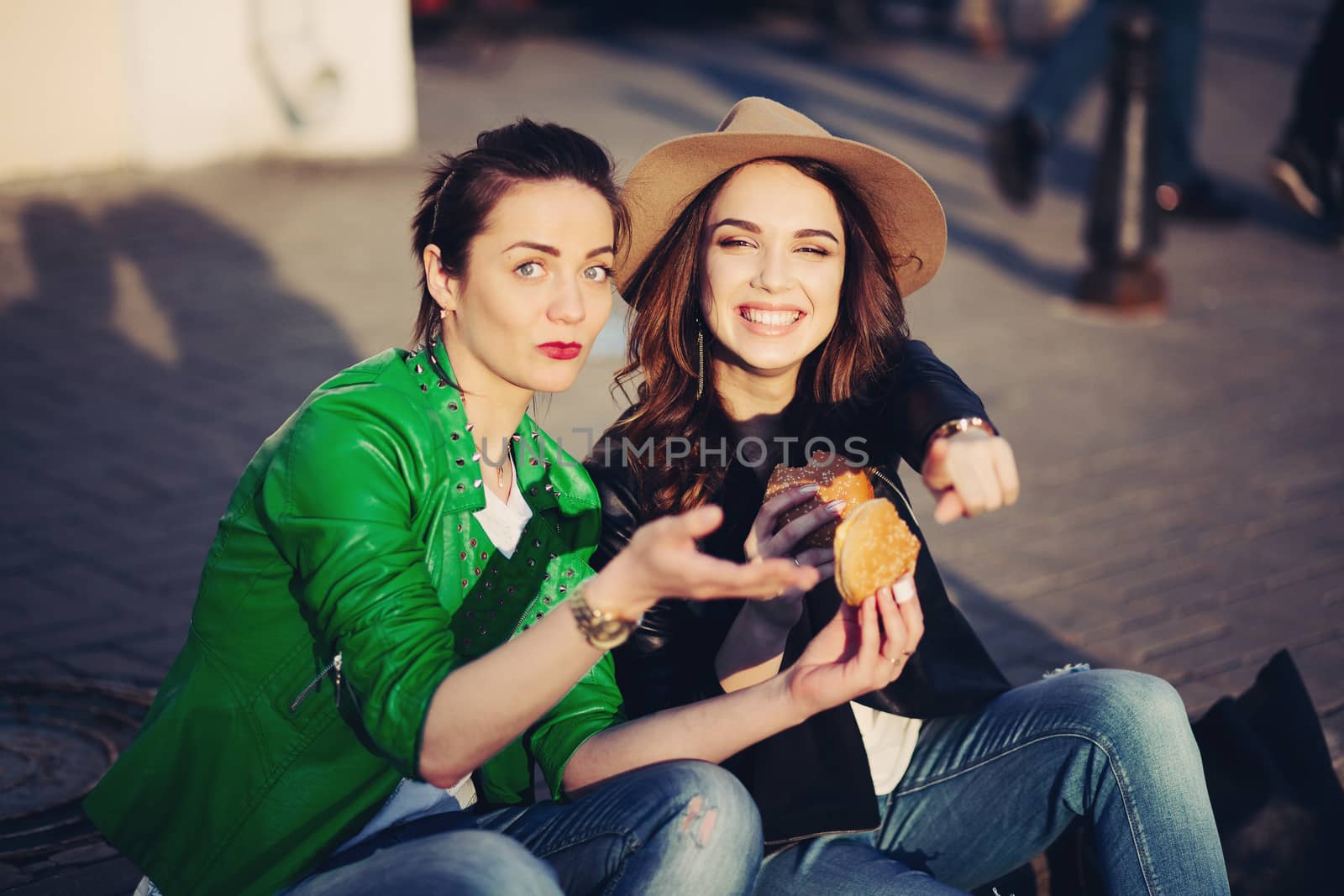 Two best friends two beautiful and stylish women, sitting at street, having dinner together, and telling each other gossip. Fashionable girls eating hamburgers and talking. Street style. Fast food.