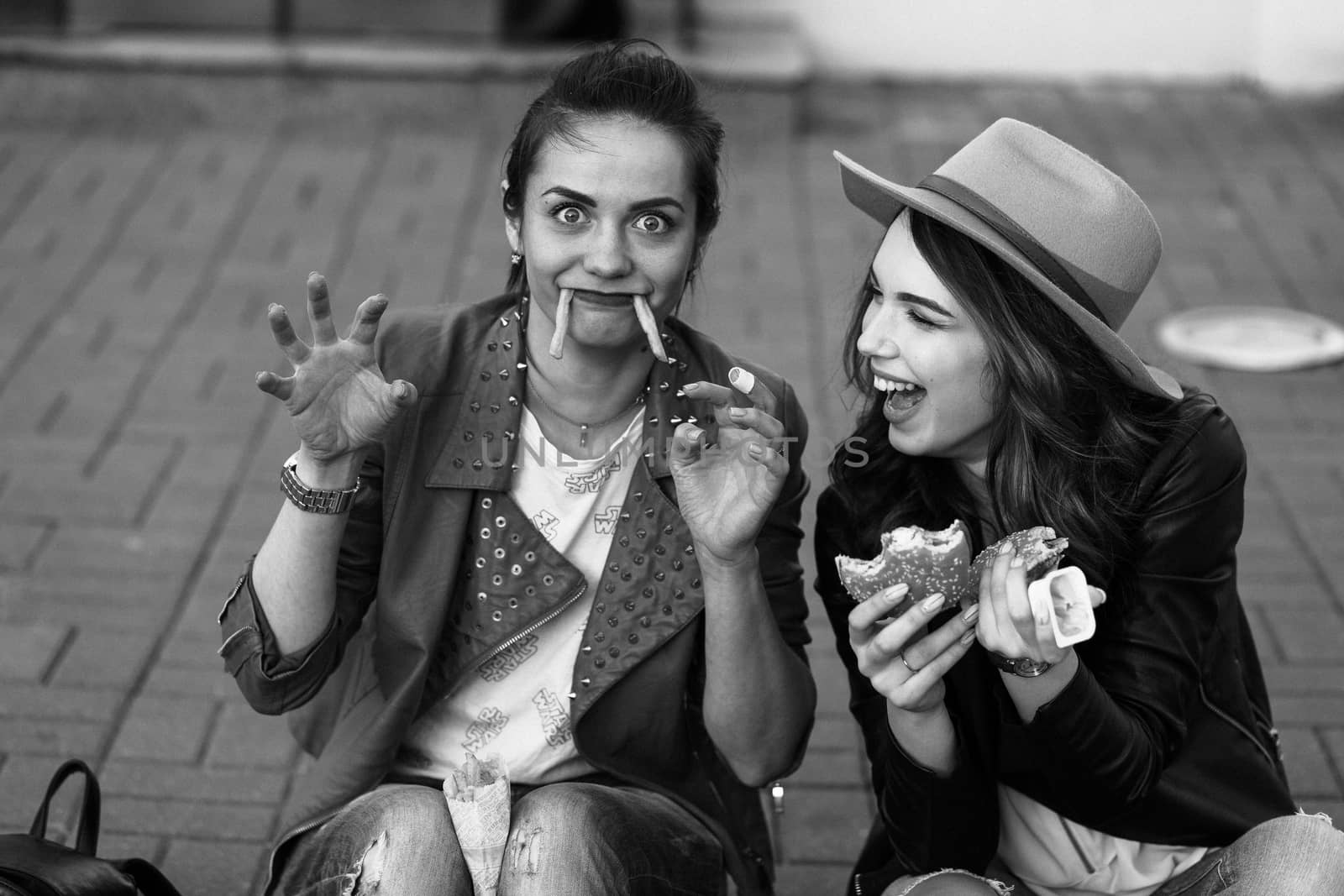 Funny girls sitting at street and eating fast food, having dinner together, and smiling to each other. Beautiful young girlfriends posing with hamburger and potato fried. Junk and unhealthy food.