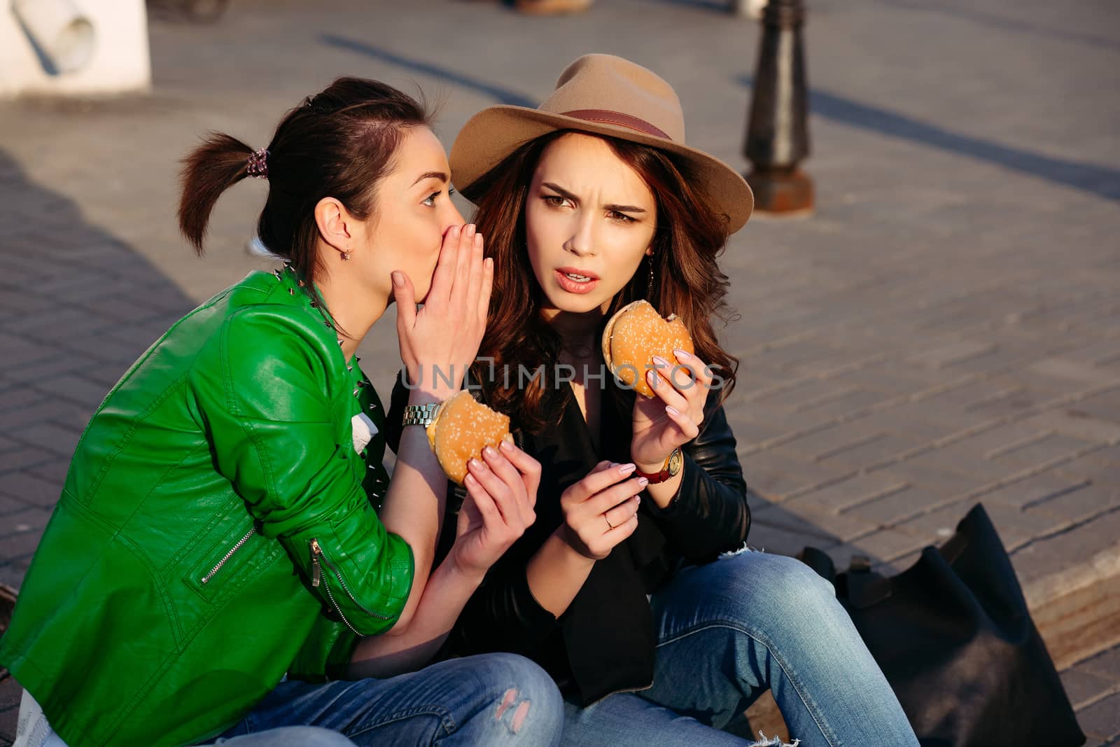 Two best friends two beautiful and stylish women, sitting at street, having dinner together, and telling each other gossip. Fashionable girls eating hamburgers and talking. Street style. Fast food.