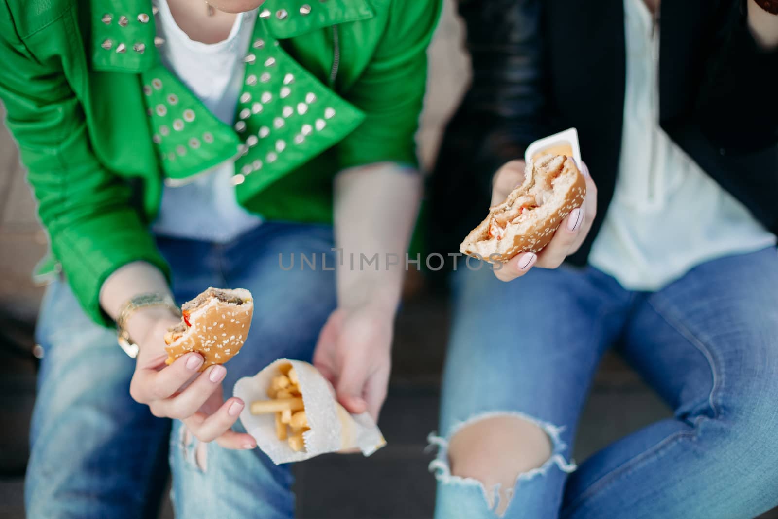 Funny girls sitting at street and eating fast food, having dinner together, and smiling to each other. Beautiful young girlfriends posing with hamburger and potato fried. Junk and unhealthy food.