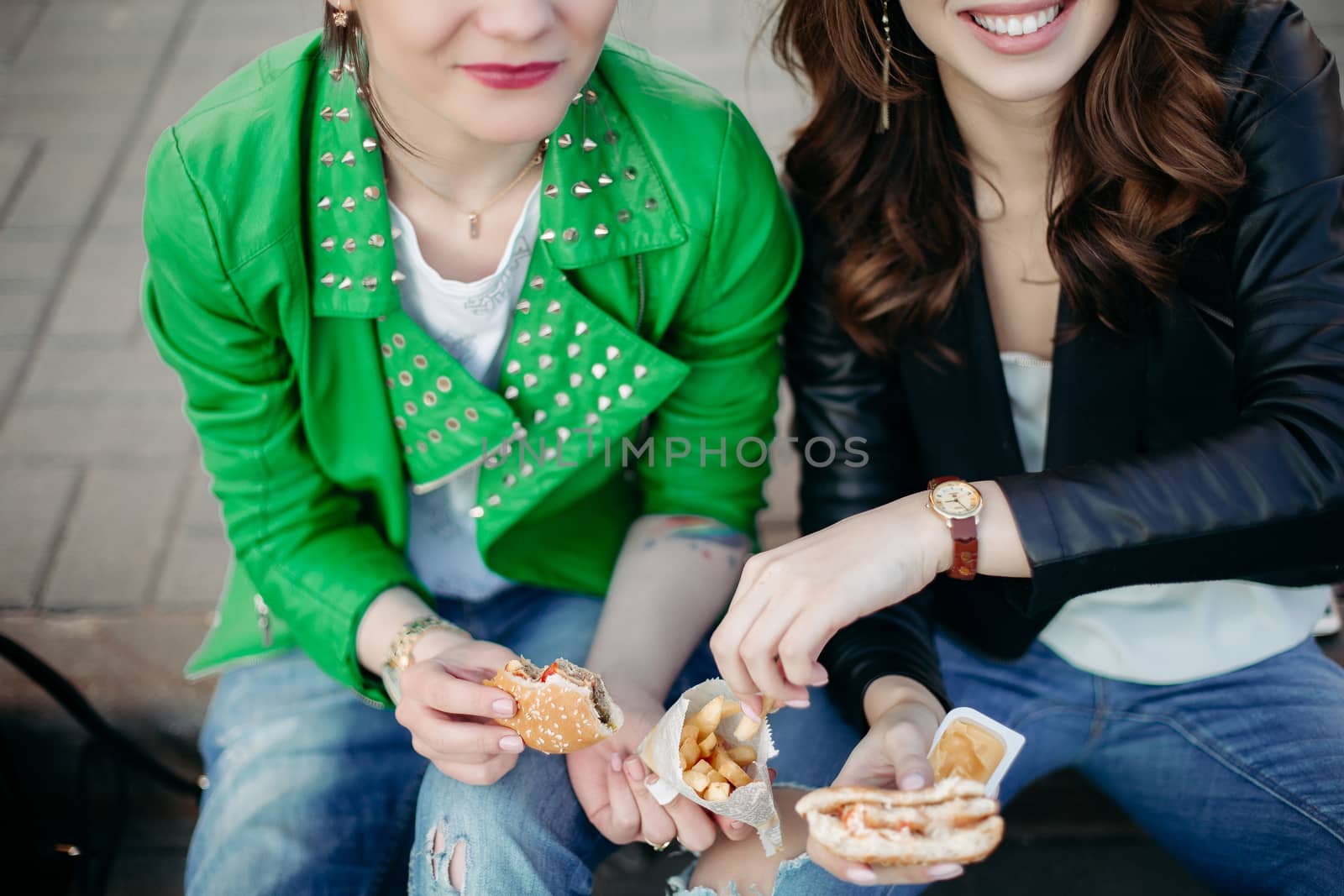 Funny girls sitting at street and eating fast food, having dinner together, and smiling to each other. Beautiful young girlfriends posing with hamburger and potato fried. Junk and unhealthy food.