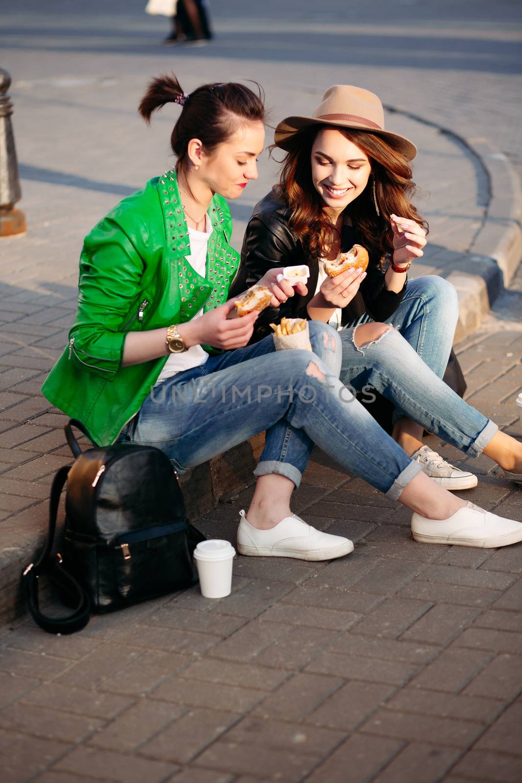 Funny girls sitting at street and eating fast food, having dinner together, and smiling to each other. Beautiful young girlfriends posing with hamburger and potato fried. Junk and unhealthy food.