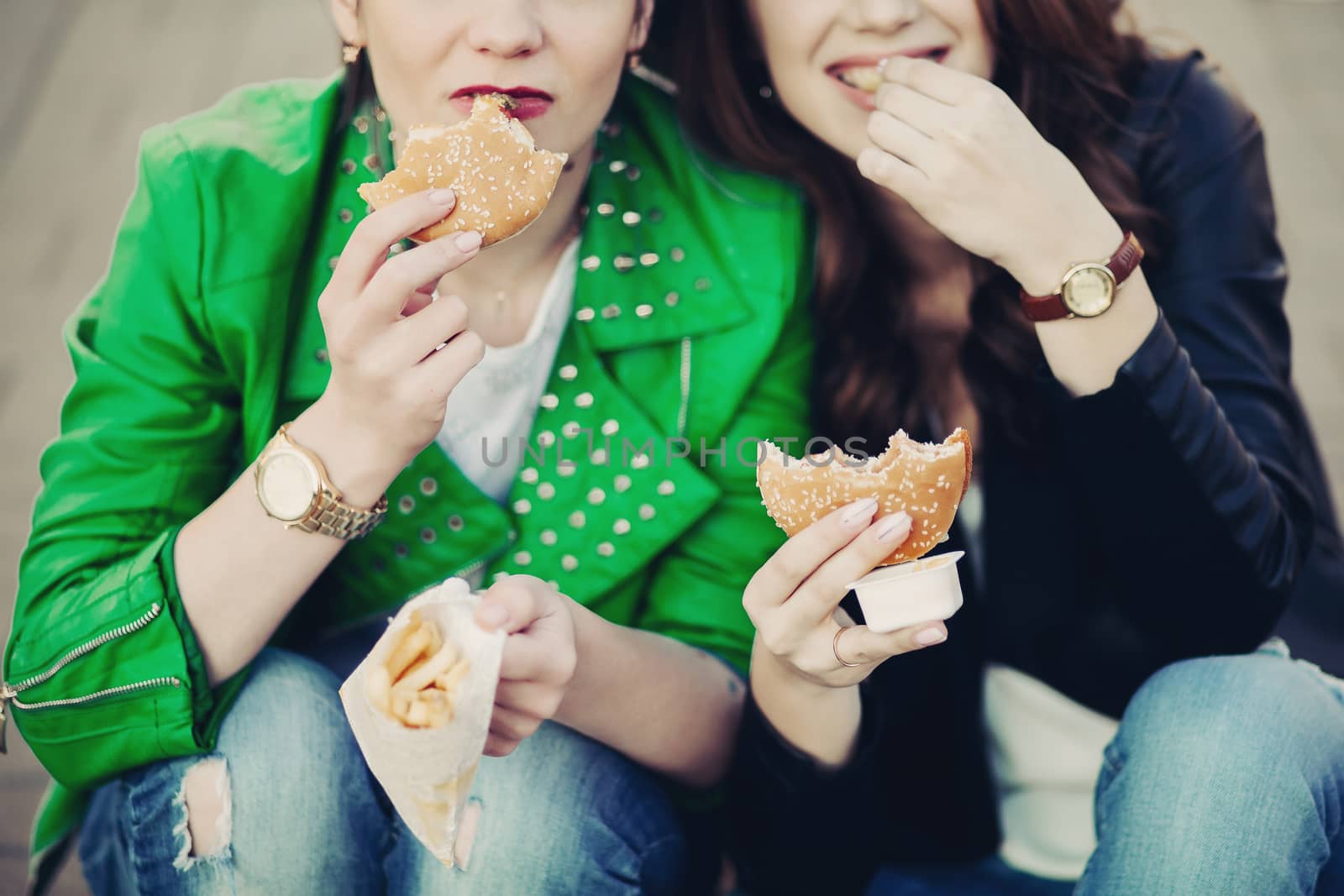 Funny girls sitting at street and eating fast food, having dinner together, and smiling to each other. Beautiful young girlfriends posing with hamburger and potato fried. Junk and unhealthy food.