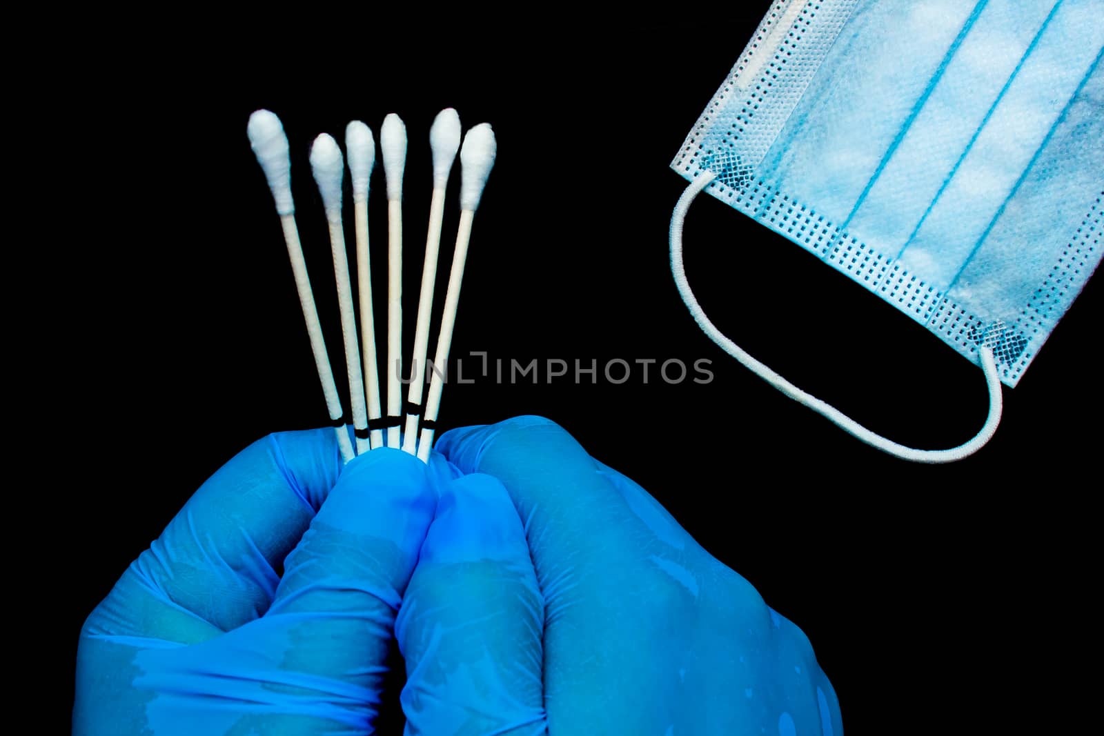 A close up of hands wearing gloves to a medical personal holding throat swabs next to a medical face mask on a black background.