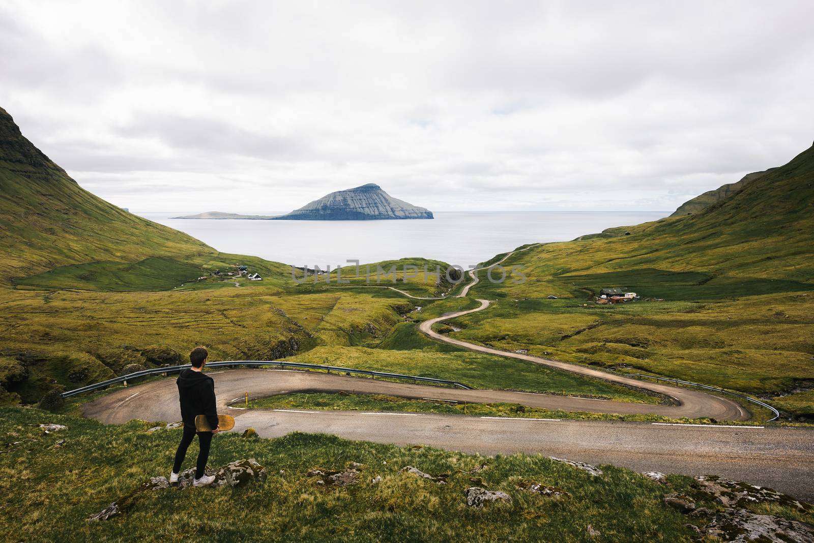 Young skater with his skateboard looks at a winding road on Faroe Islands surrounded by beatiful scenery and Atlantic Ocean in the background