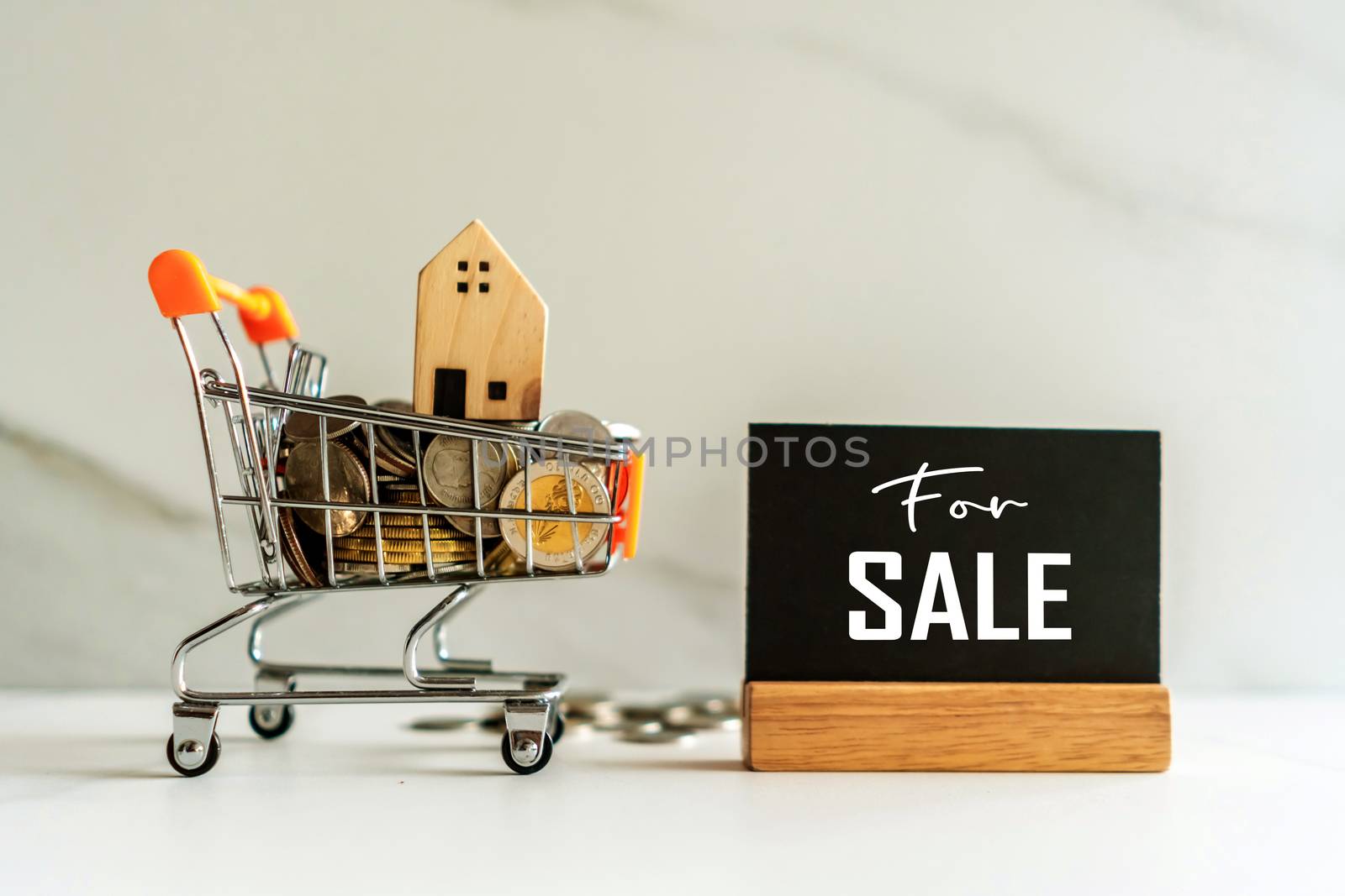 Home model in mini cart model full of coins money with for sale text on blank black wooden sign background.