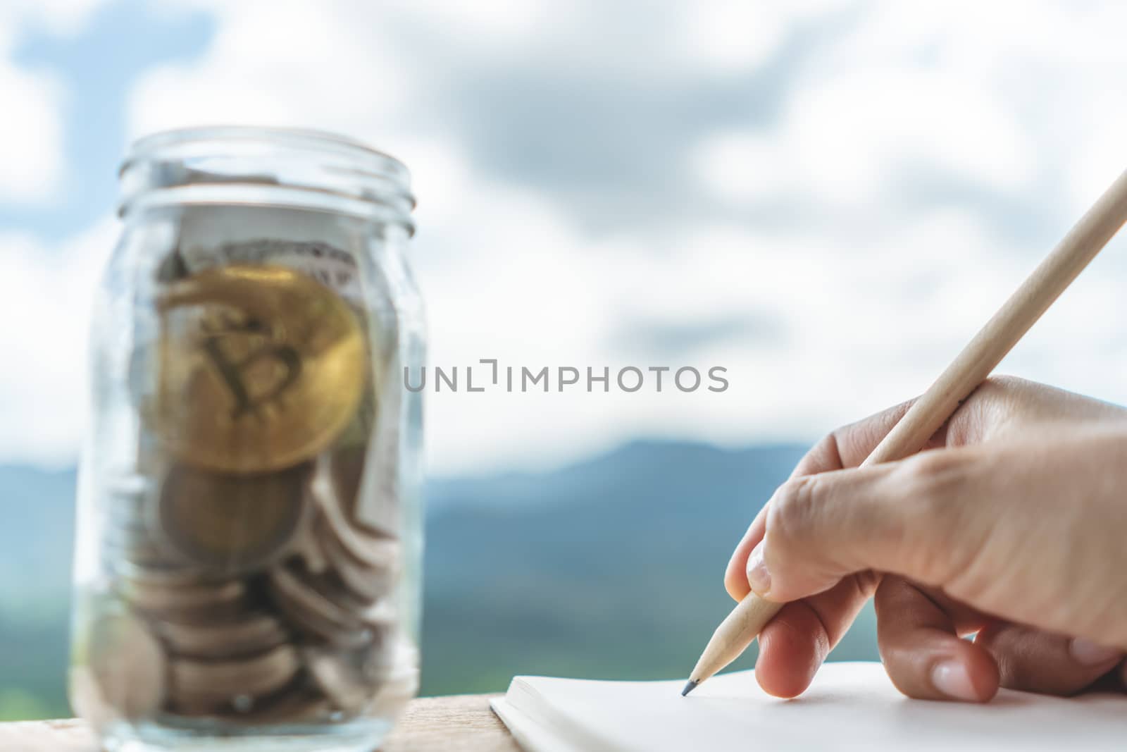 Woman hand writing down in small white memo notebook with jar full of money for plan wealth investing for future. by Suwant