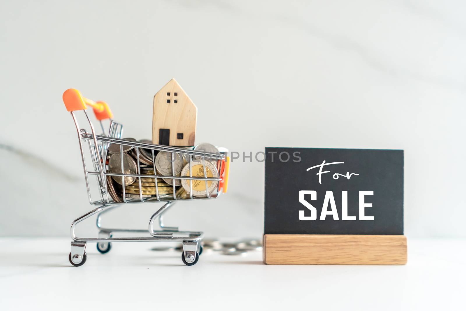 Home model in mini cart model full of coins money with for sale text on blank black wooden sign background.