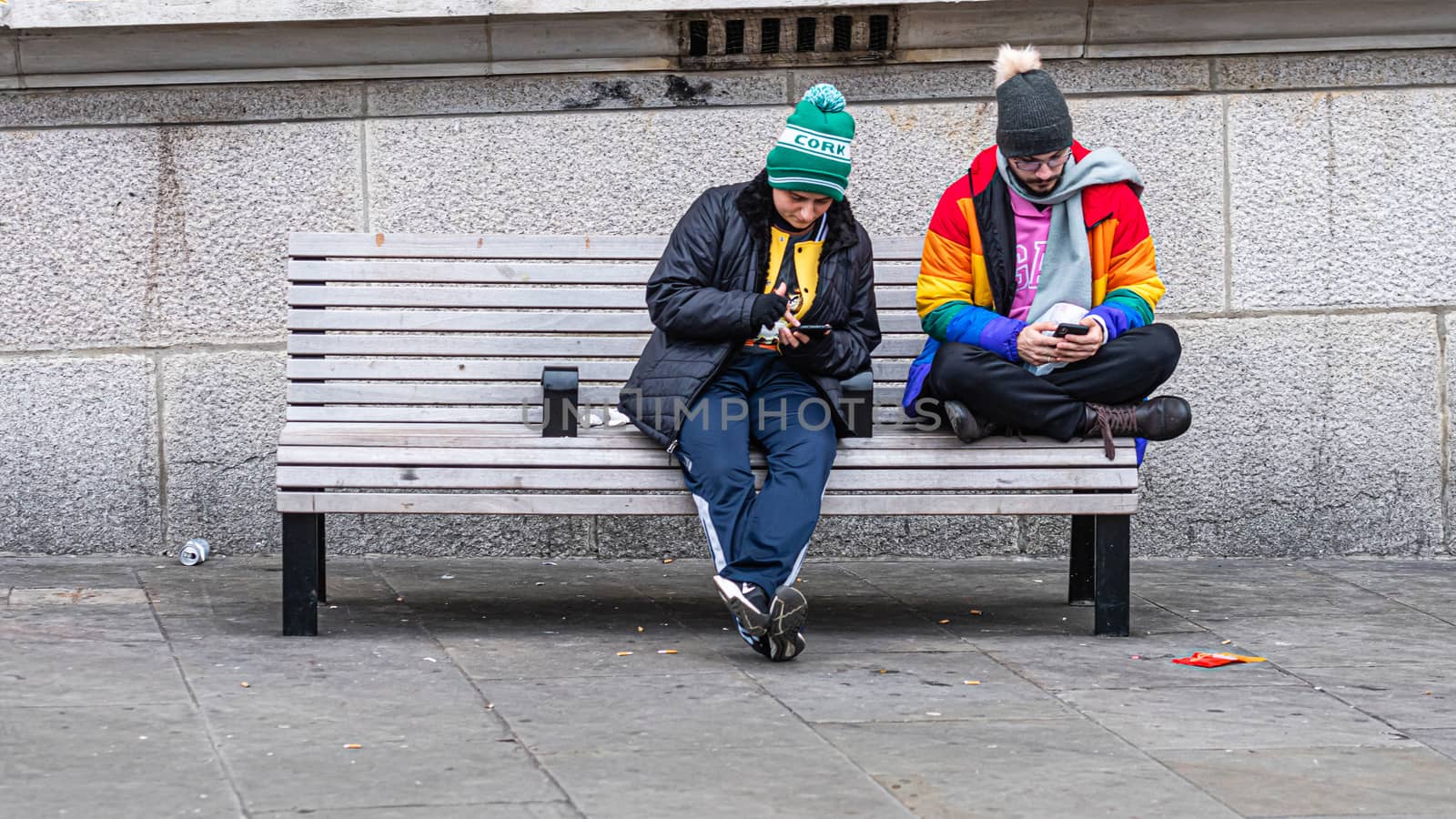 London, UK - January 1, 2020: Two men sit on a bench on the side of the street and look at their smartphones