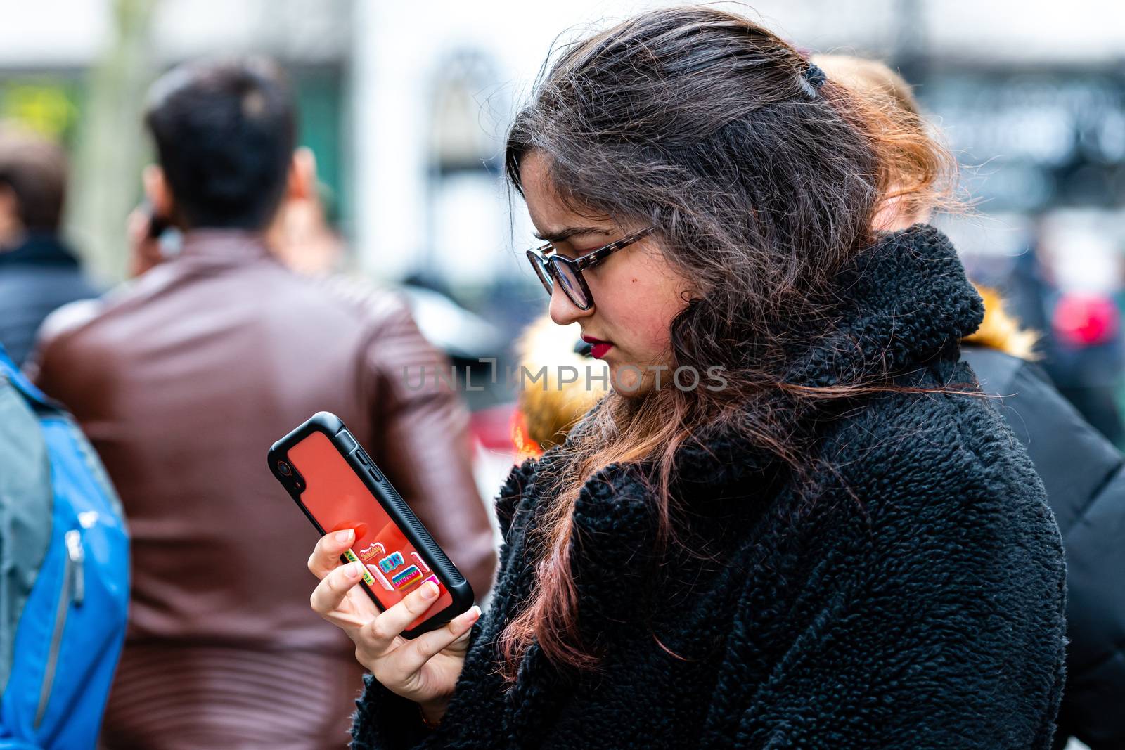 London, England, UK - January 2, 2020: Young woman with glasses standing in crowd of people staring at smartphone