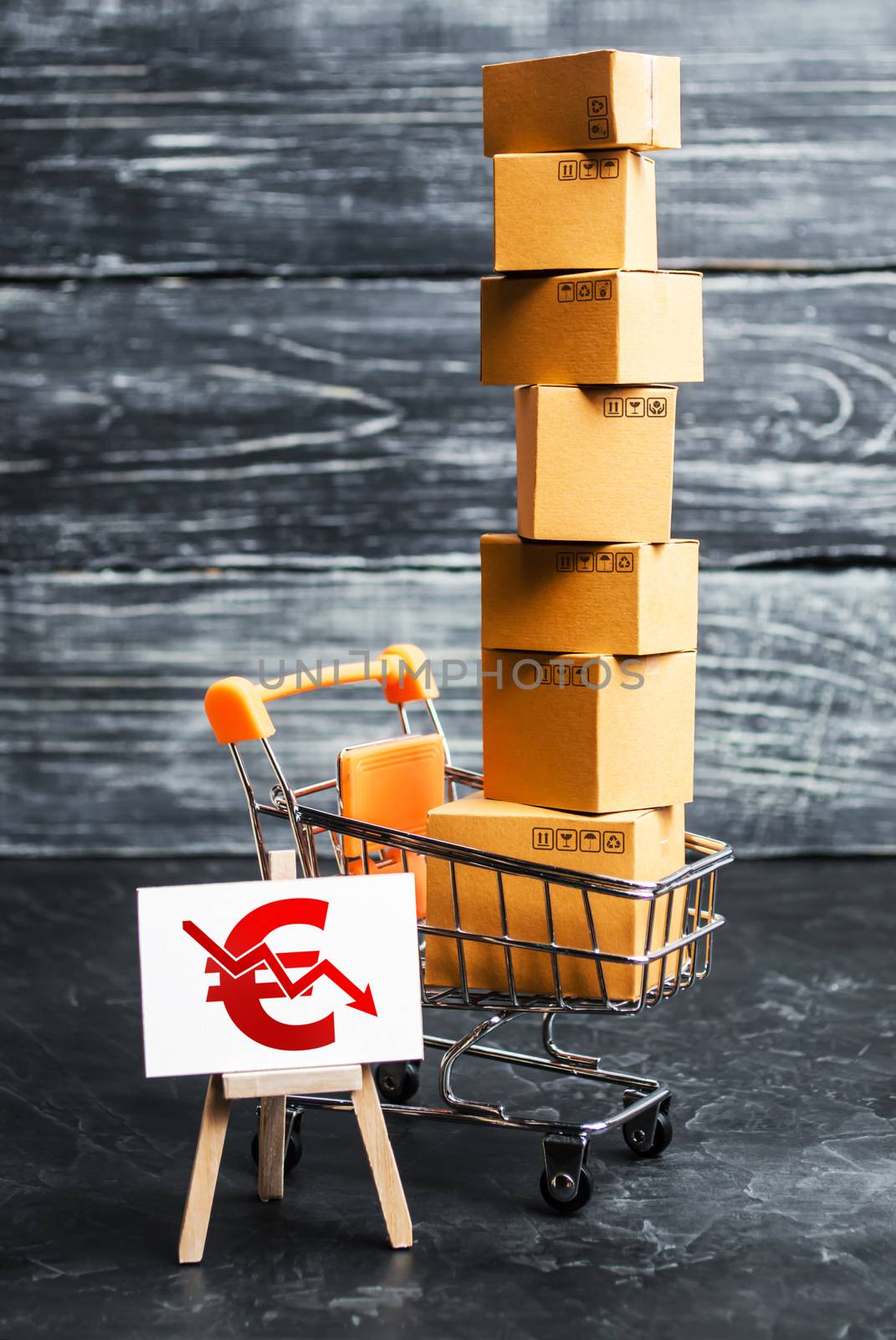 A shopping cart loaded with boxes and a sign with a euro symbol down arrow. Decrease in income from sales of goods and retail. Fall in purchasing power. Price reduction, discounts by iLixe48