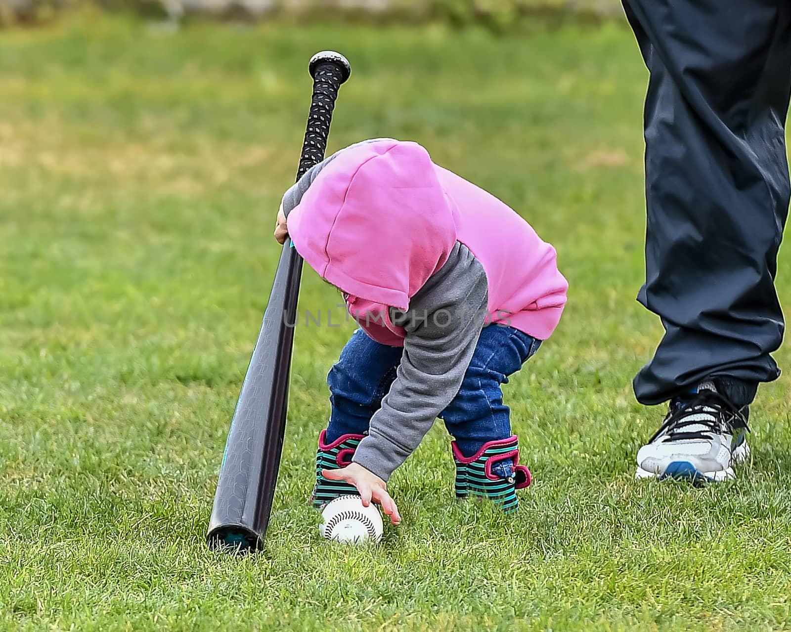 Adorable infant playing with a baseball bat and ball at the baseball park. Cute baby with headband trying to pick up the baseball and also trying to hit the ball with a bat that is too big for her.