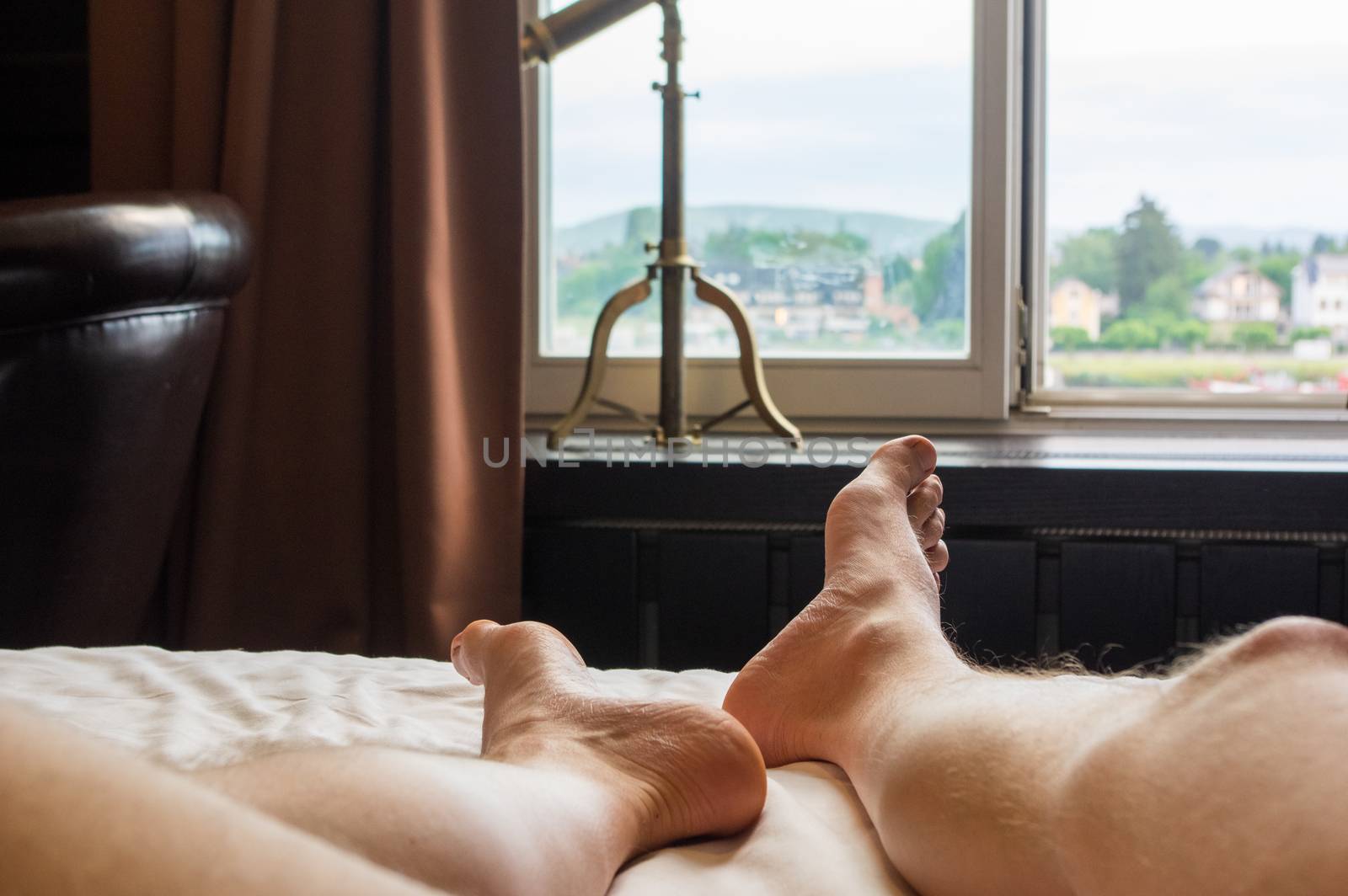 Legs and feet of a caucasian man lying on his back in a hotel room looking out of a window by MarcoWarm