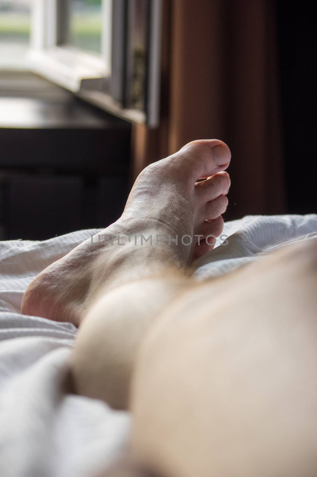 Close-up of male leg and foot lying on a bed in a hotel room looking out of a window by MarcoWarm