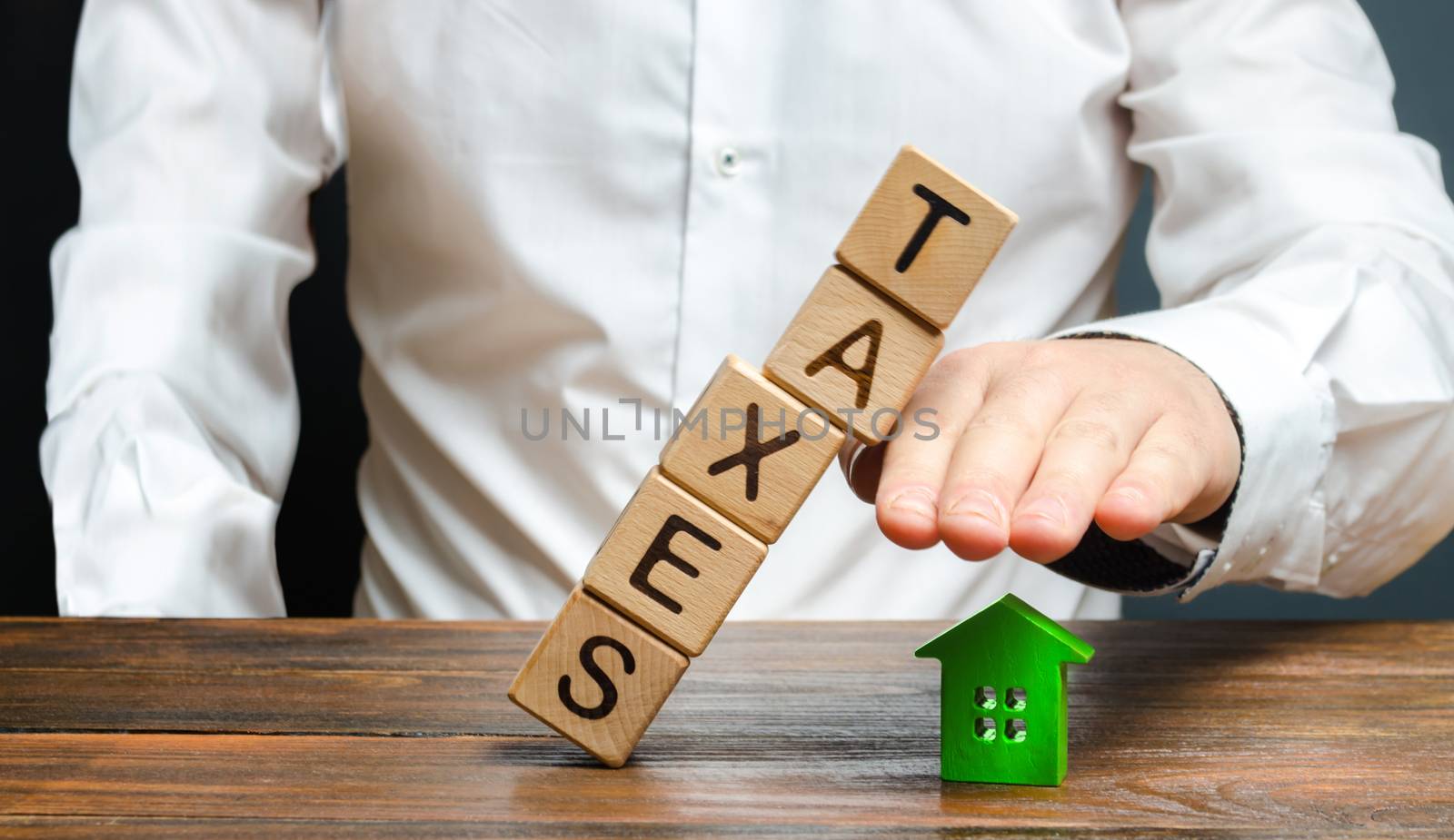 A man protects his hand with a figurine of a house from a falling tower of cubes with the word taxes. Heavy tax burden, protecting the interests of small and medium-sized businesses, competitiveness. by iLixe48