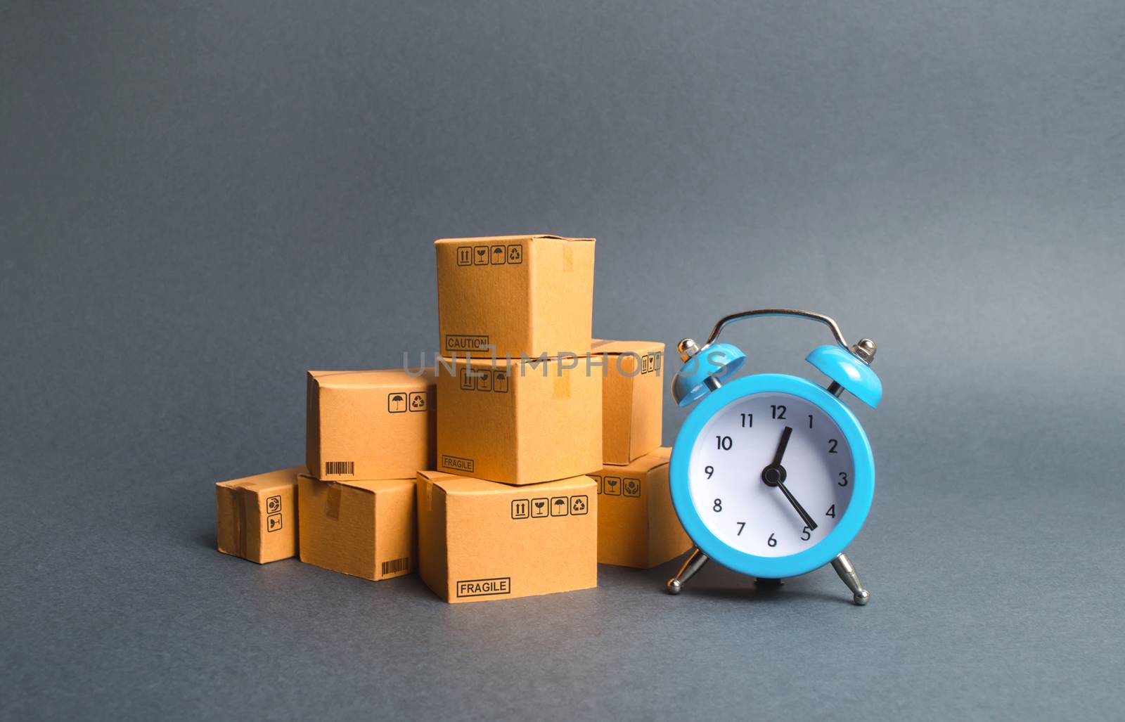 A stack of cardboard boxes and a blue alarm clock. Express delivery concept. Temporary storage, limited offer and discount. Optimization of logistics and delivery, improving efficiency, reducing costs by iLixe48