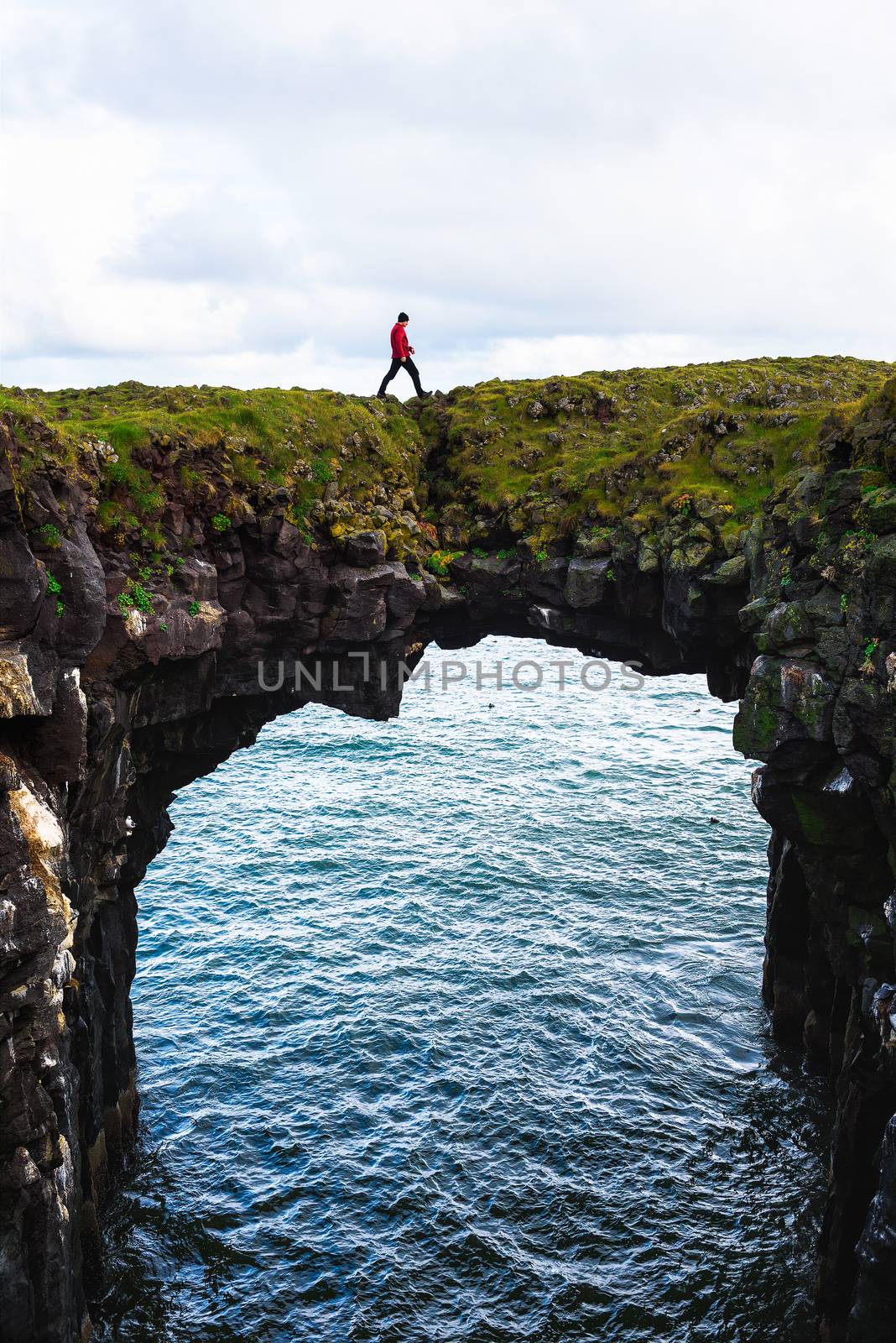 Tourist walks over a natural rock bridge connecting basalt cliffs in Arnarstapi, Iceland, with Atlantic Ocean in the background.