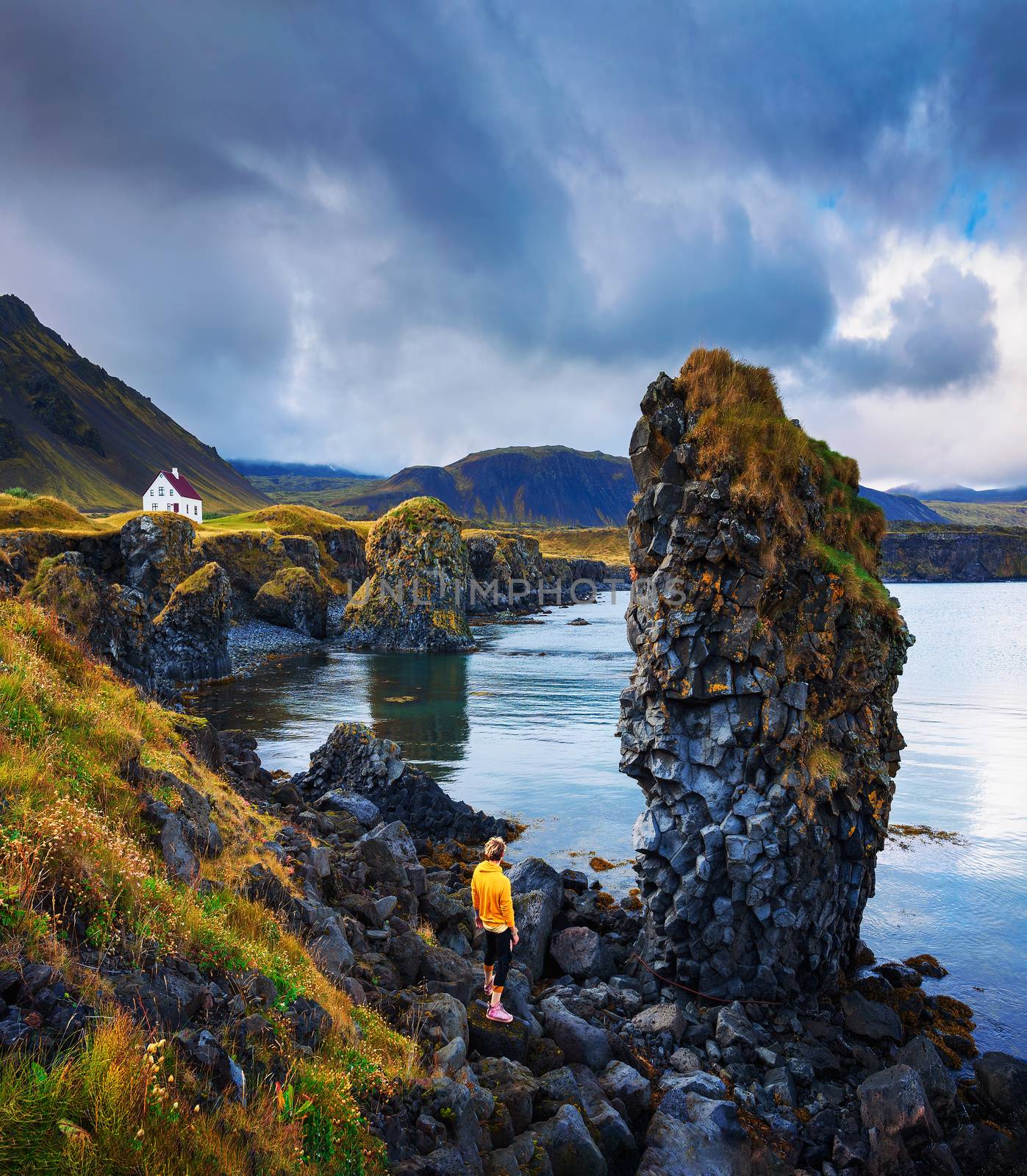 Tourist stands on a rocky beach of Snaefellsnes peninsula in Iceland under a big cliff, and looks at a small white house in the village of Arnarstapi.