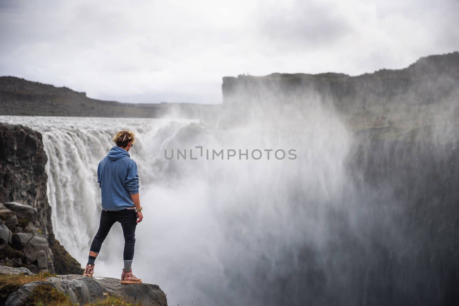 Young hiker standing at the edge of the Dettifoss waterfall located on the Jokulsa a Fjollum river in Iceland. Dettifoss is the second most powerful waterfall in Europe after the Rhine Falls.