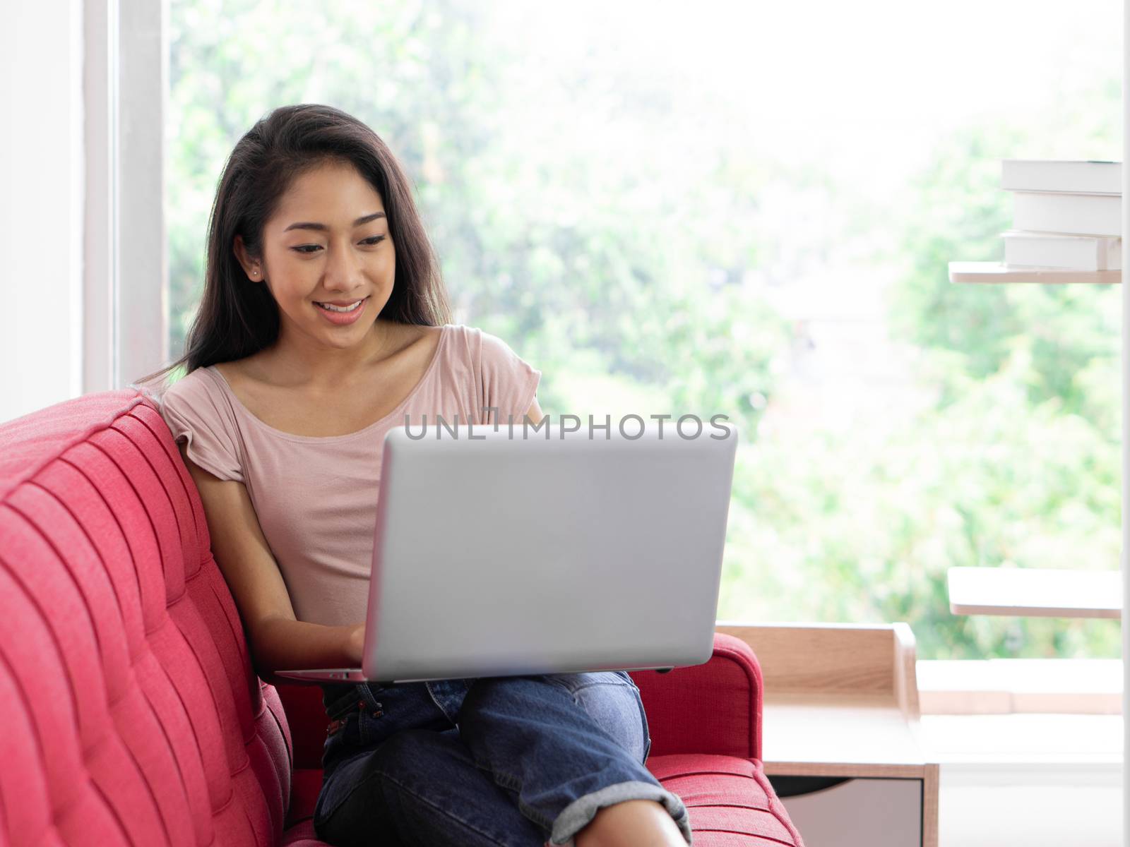 Young asian teen use laptop computer on the red sofa to relax in the living room, enjoy leisure weekend at home. Stress free concept
