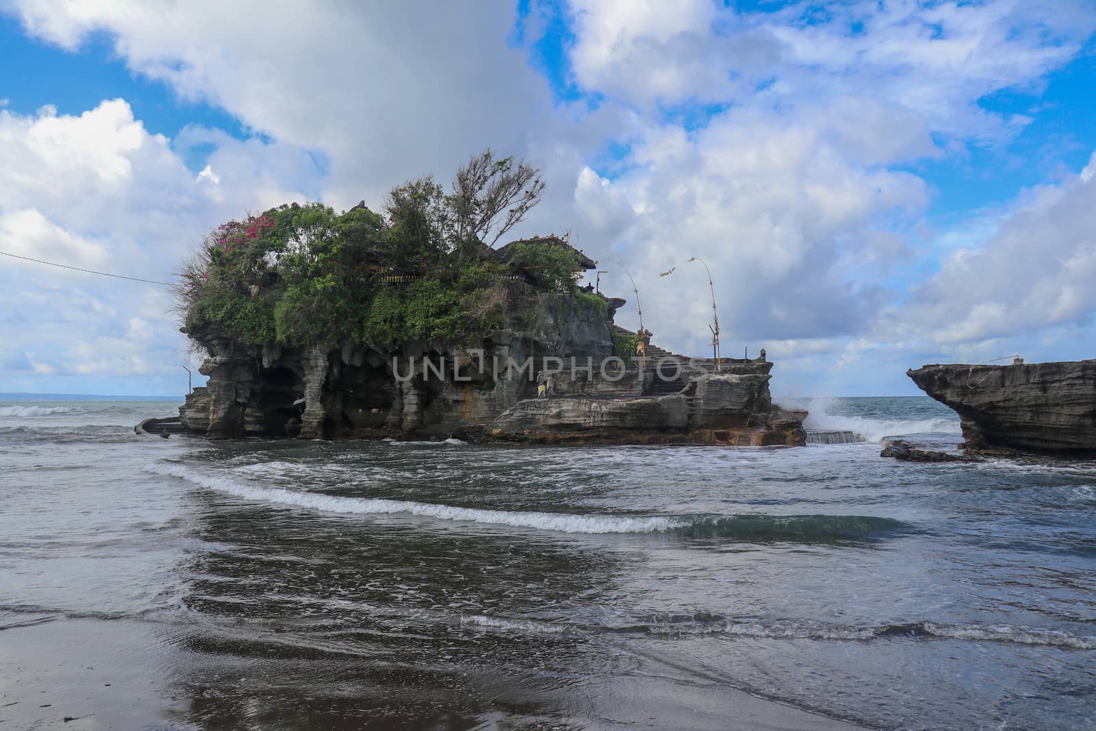 Waves shatter on a cliff at the top of which is the Hindu temple of Tanah Lot. Temple built on a rock in the sea off the coast of Bali island, Indonesia. Lush tropical vegetation on top of a cliff.