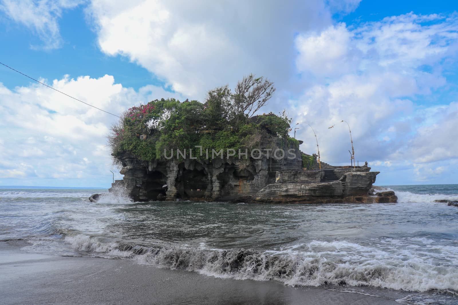 Waves shatter on a cliff at the top of which is the Hindu temple of Tanah Lot. Temple built on a rock in the sea off the coast of Bali island, Indonesia. Lush tropical vegetation on top of a cliff.