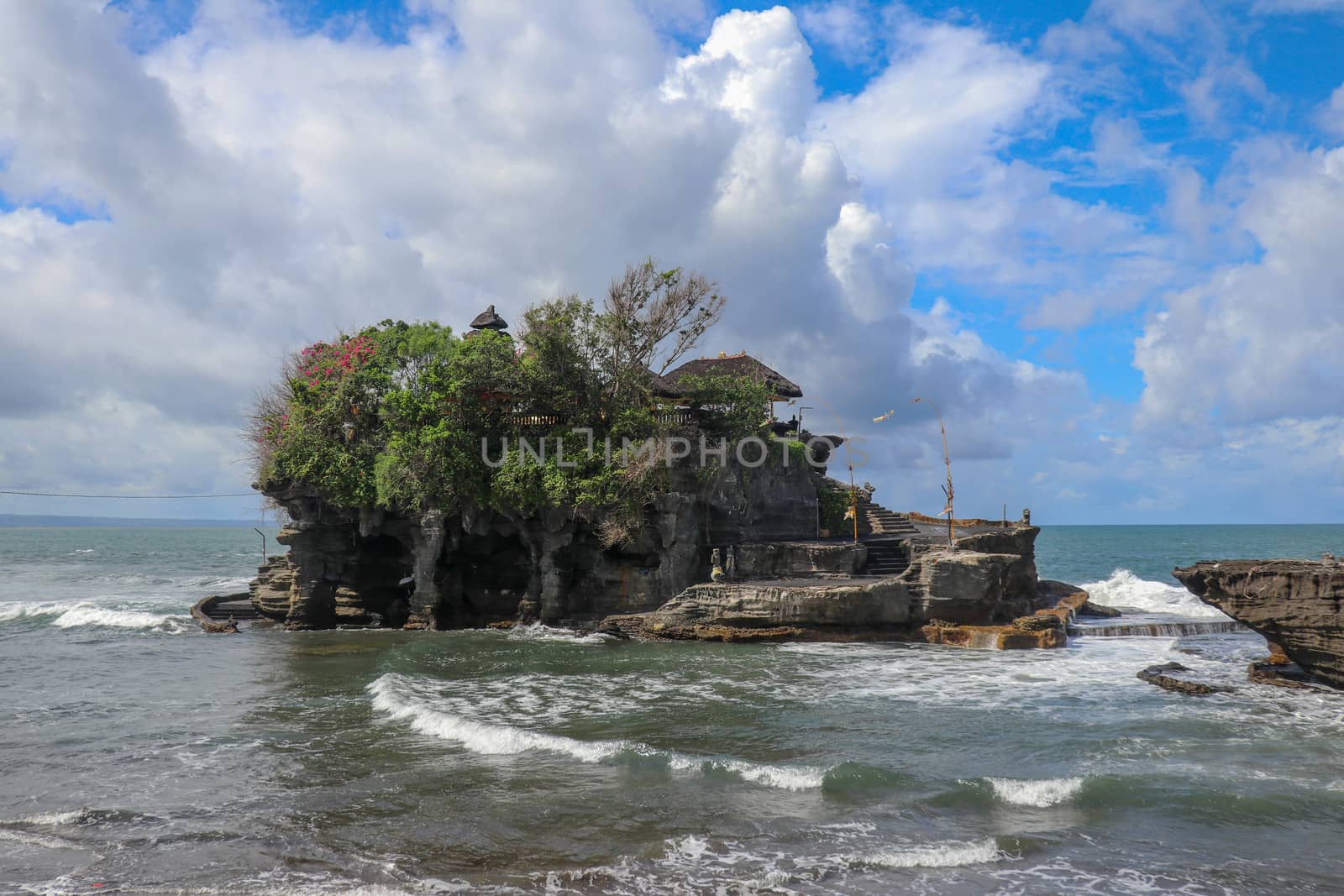 Waves shatter on a cliff at the top of which is the Hindu temple of Tanah Lot. Temple built on a rock in the sea off the coast of Bali island, Indonesia. Lush tropical vegetation on top of a cliff.