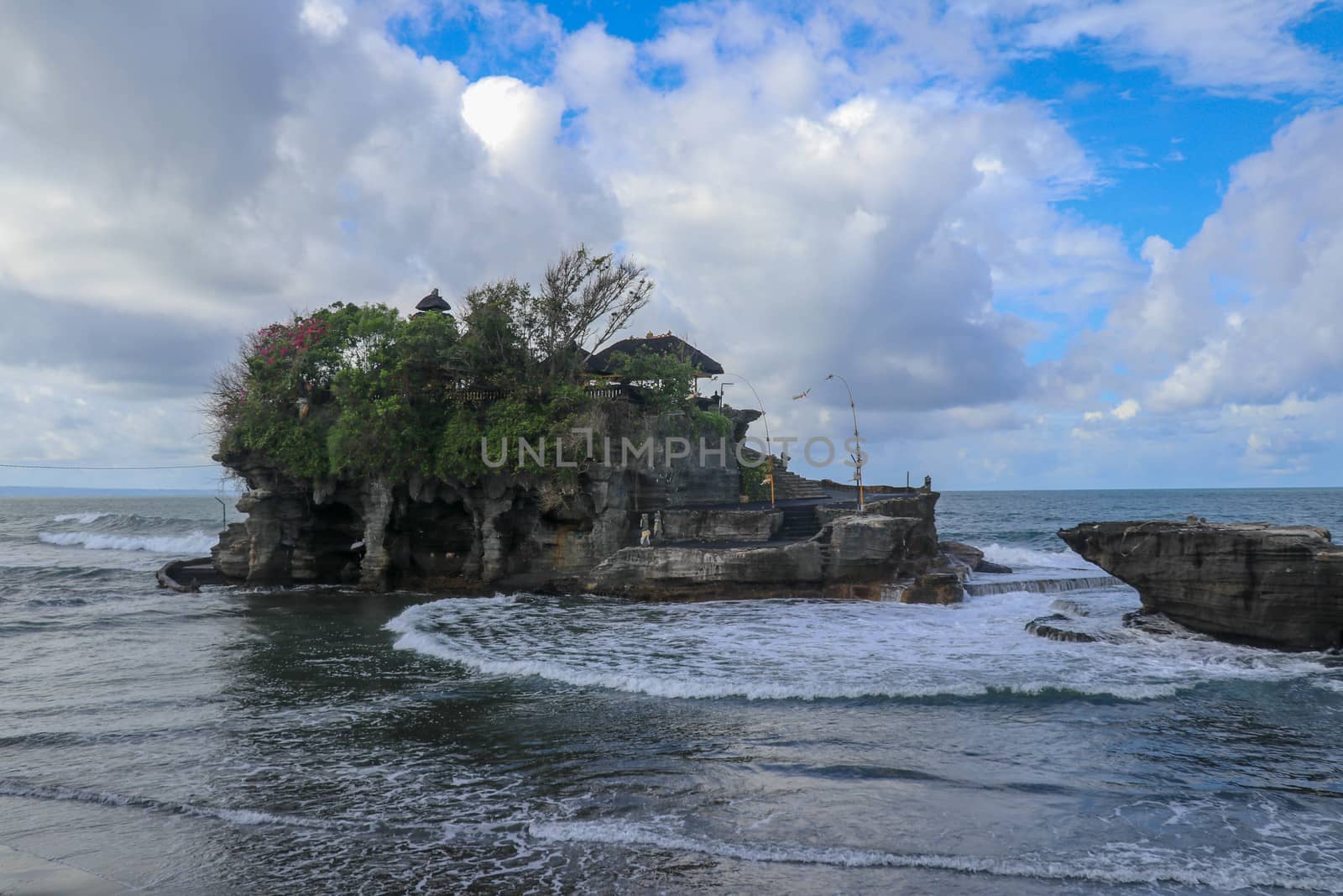 Waves shatter on a cliff at the top of which is the Hindu temple of Tanah Lot. Temple built on a rock in the sea off the coast of Bali island, Indonesia. Lush tropical vegetation on top of a cliff.