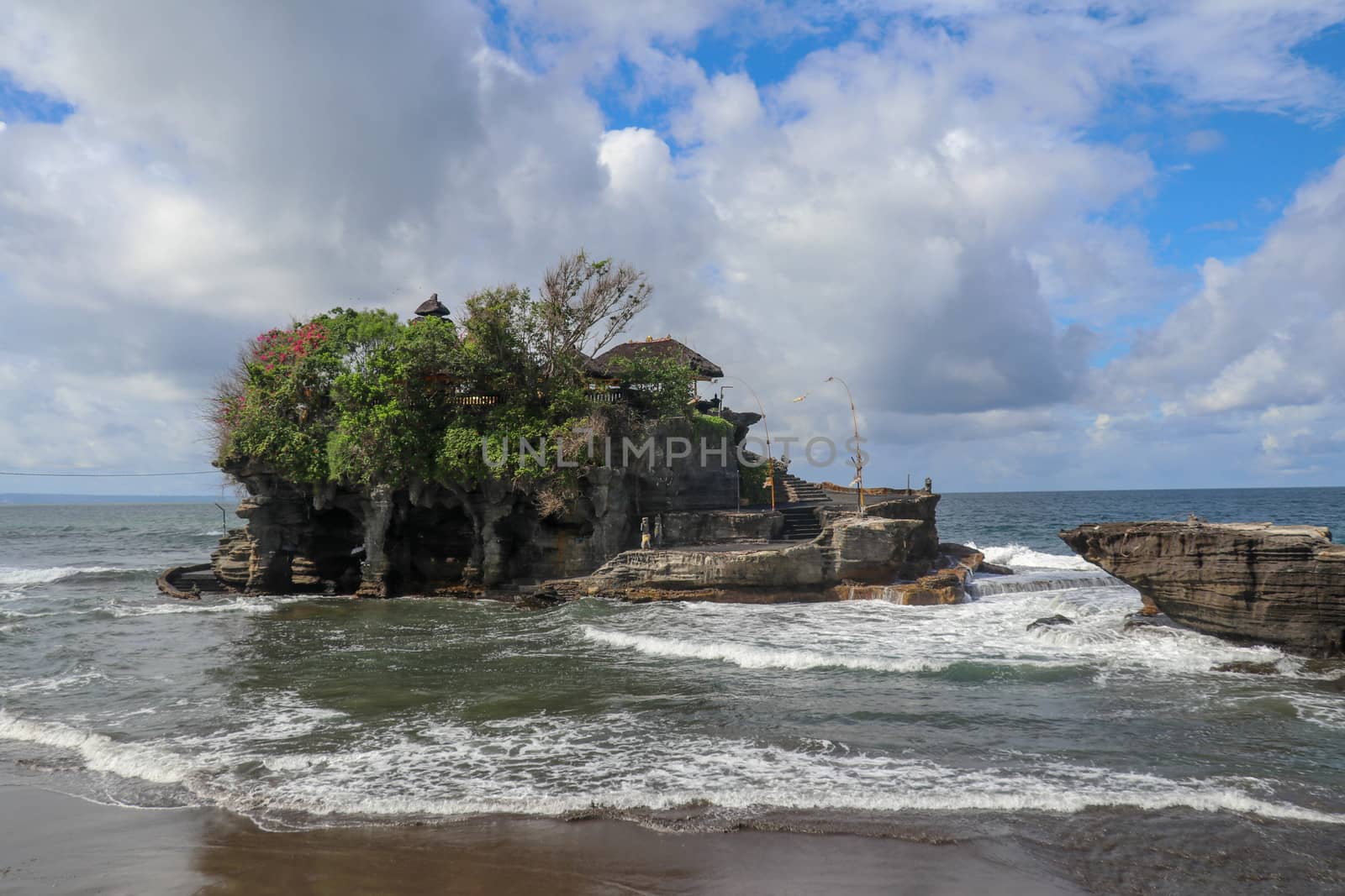 Waves shatter on a cliff at the top of which is the Hindu temple of Tanah Lot. Temple built on a rock in the sea off the coast of Bali island, Indonesia. Lush tropical vegetation on top of a cliff.