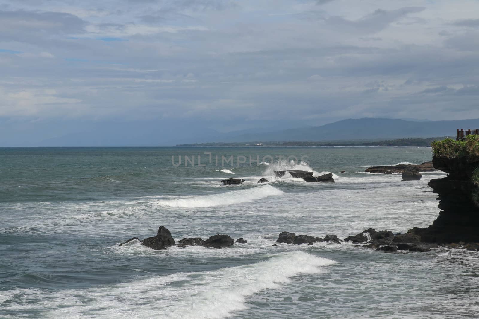 Rock cliffs in the Indian Ocean off the coast of Bali island, near Batu Bolong and tourism attraction spot Tanah Lot temple. Waves are shattering on rocks of solidified black volcanic lava. Indonesia.